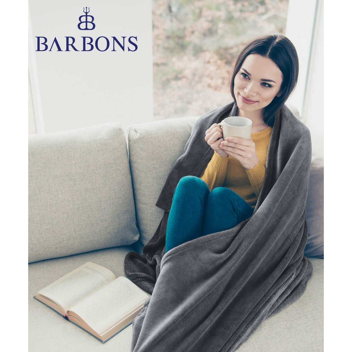 Eine Frau, die sich in eine graue Decke auf einem Sofa hüllt, hält eine Tasse und ein Buch.