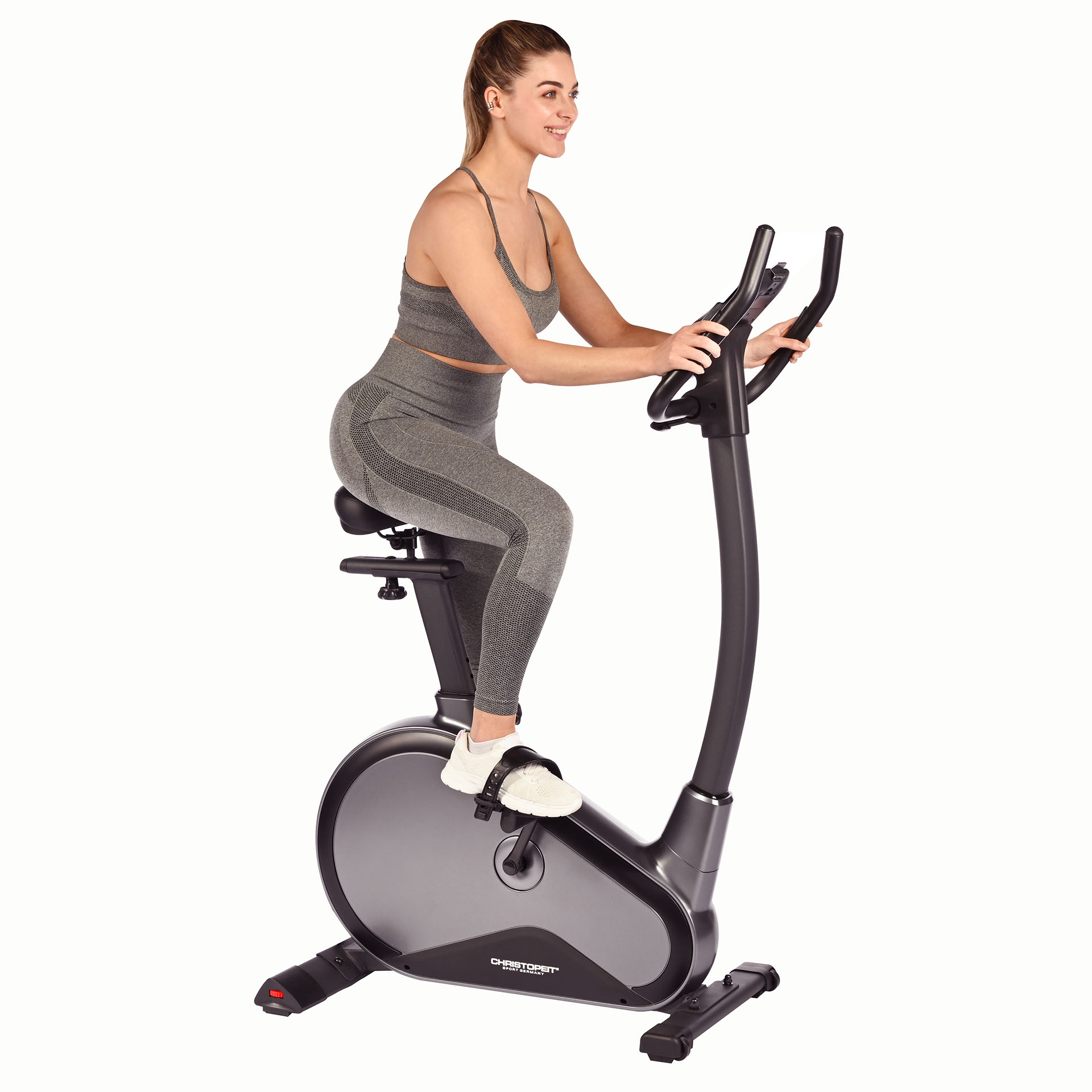 Eine Frau in grauer Sportkleidung fährt auf einem Heimtrainer. Weißer Hintergrund.