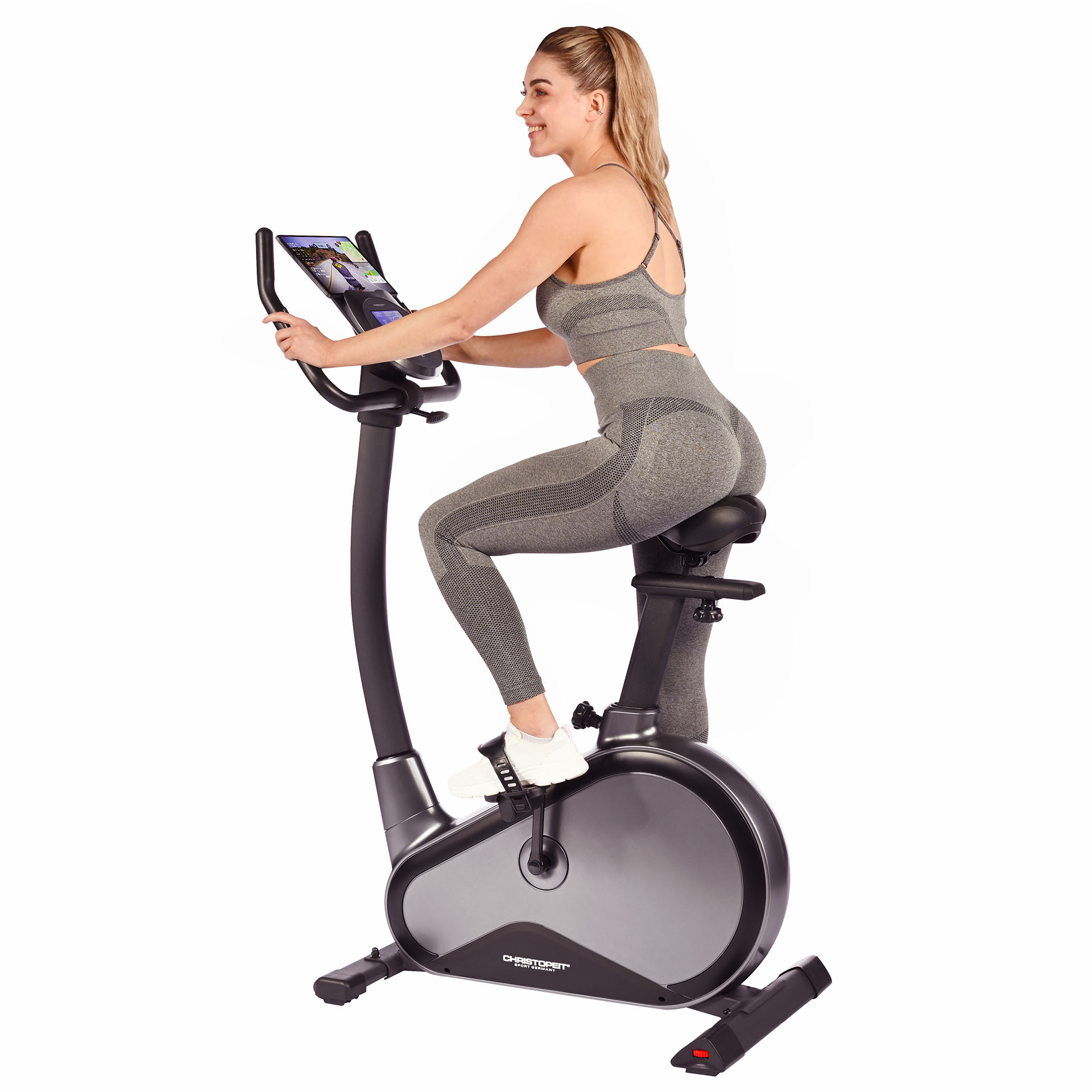 Frau in grauer Sportkleidung trainiert auf einem Heimtrainer, lächelt und schaut nach vorne.