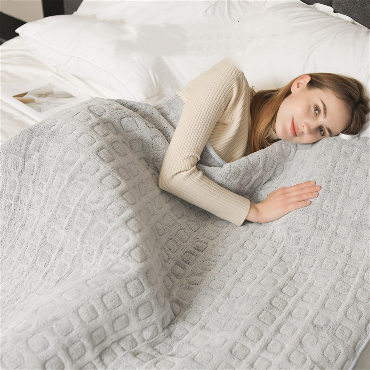 Frau liegt auf einem Bett, bedeckt mit einer grauen, gemusterten Decke, blickt in die Kamera.