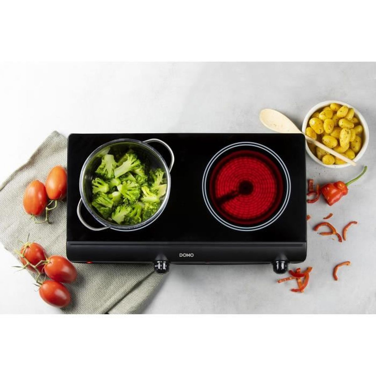 Eine schwarze Doppelkochplatte mit zwei roten Heizelementen. Brokkoli und Tomaten sind in der Nähe.