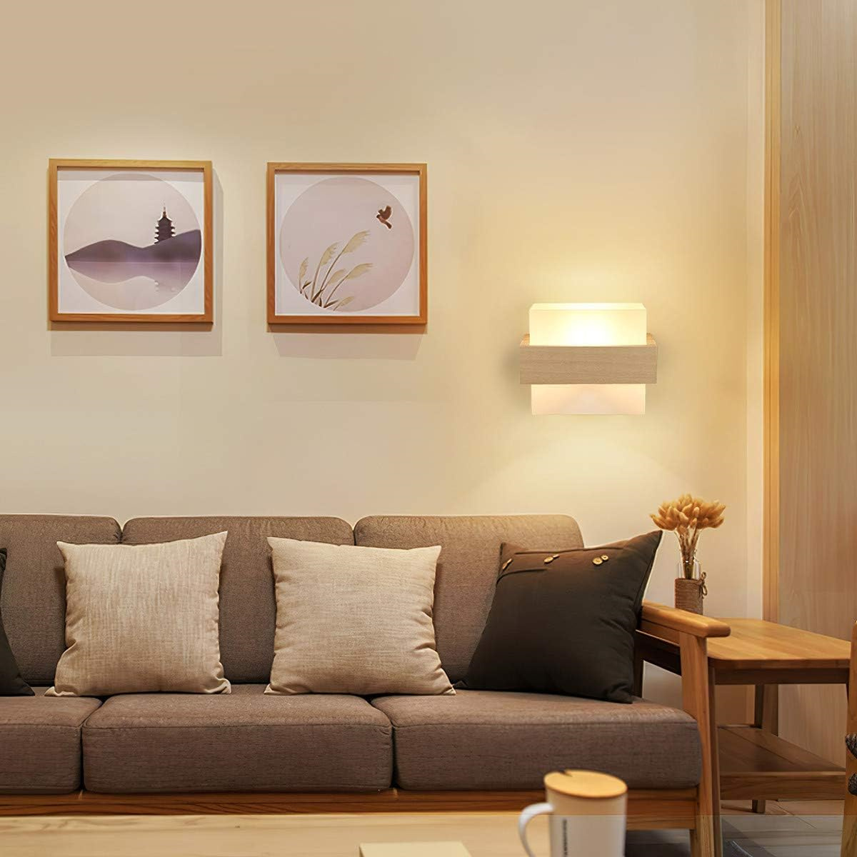 Ein Sofa mit Kissen unter zwei gerahmten Bildern und einer Wandlampe. Eine Kaffeetasse steht auf einem kleinen Tisch.