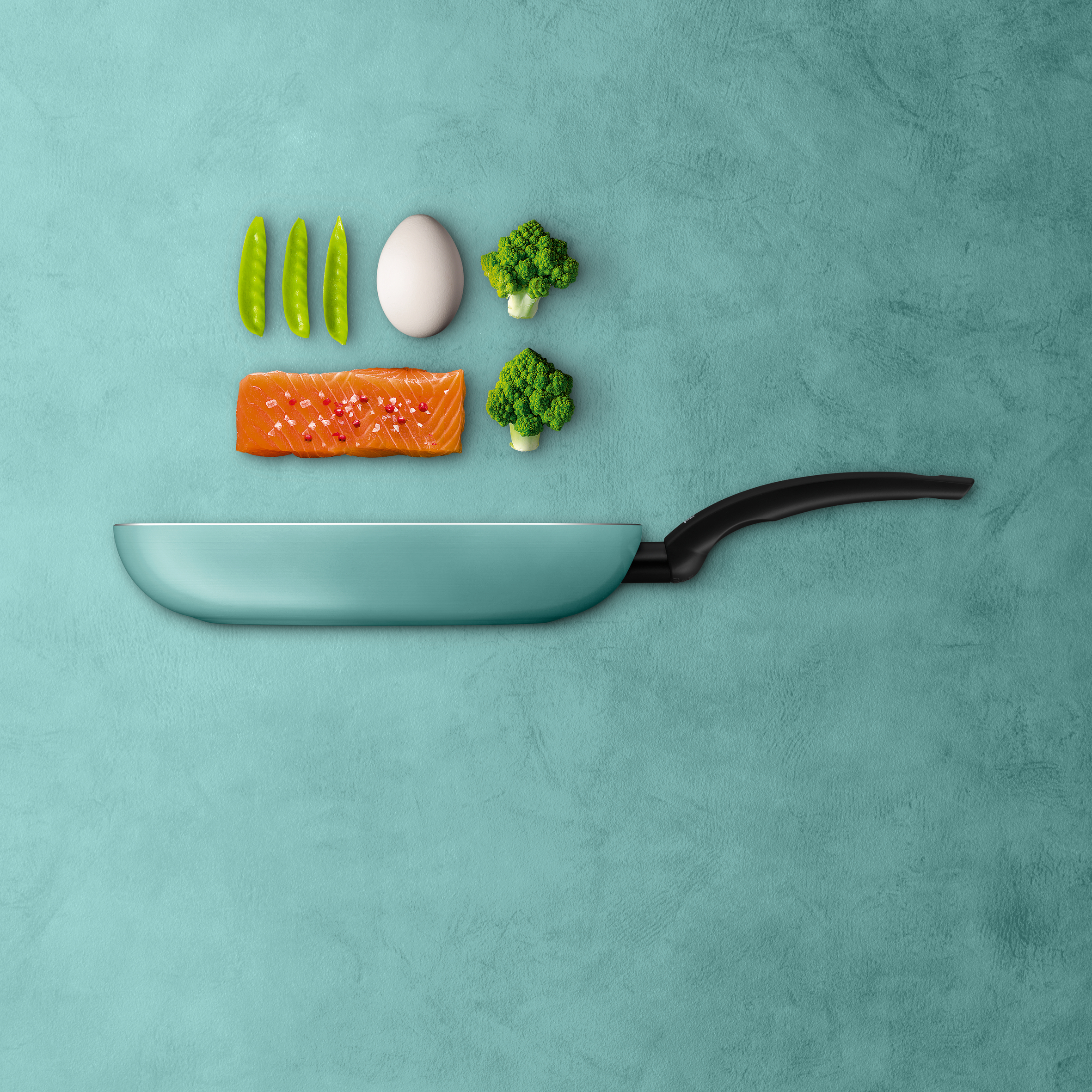 Pfanne mit Zutaten: Lachs, Ei, Erbsen, Brokkoli, auf türkisfarbenem Hintergrund.