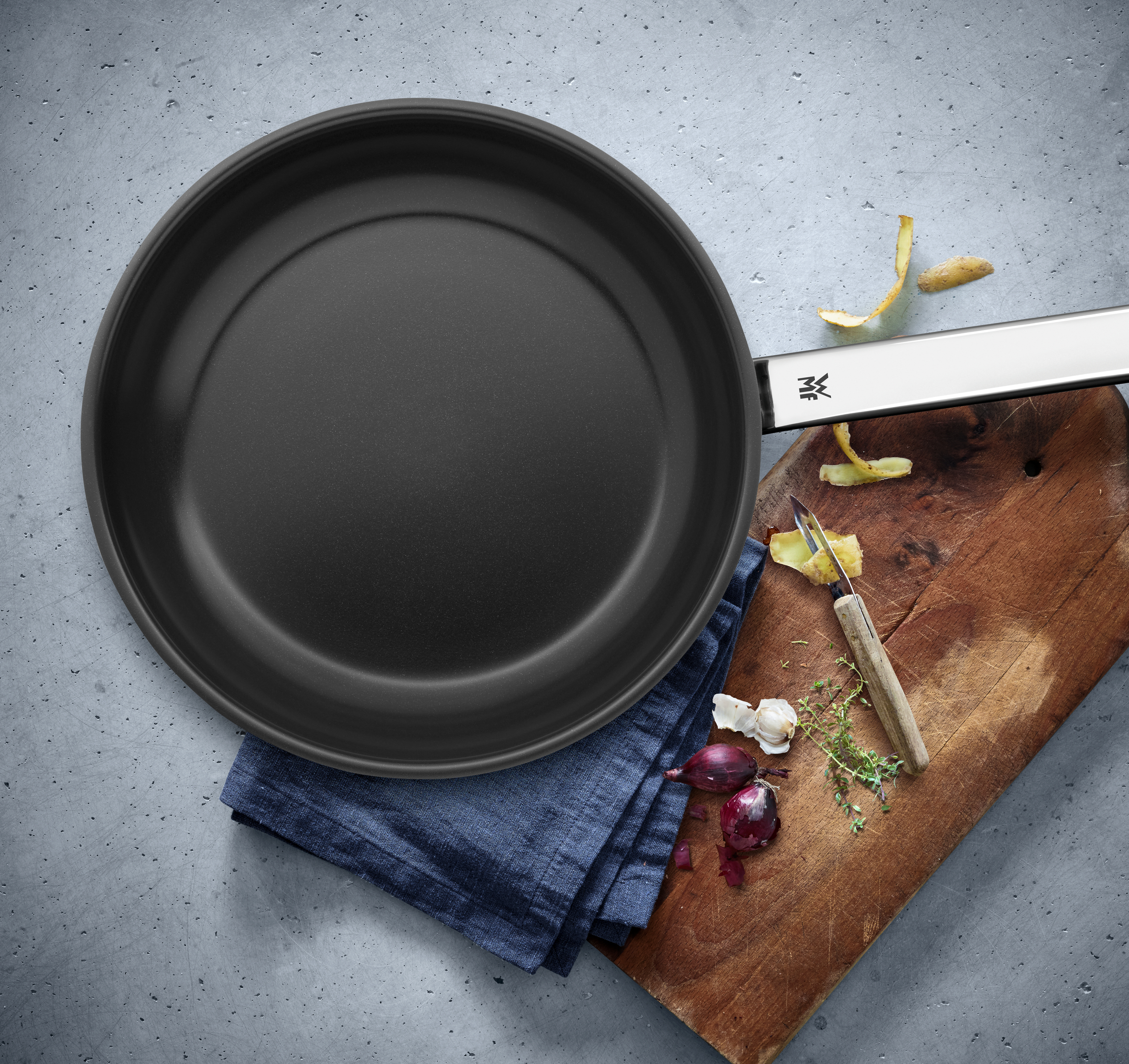 Eine schwarze Bratpfanne auf einem blauen Tuch, mit einem Holzbrett und etwas Gemüse.