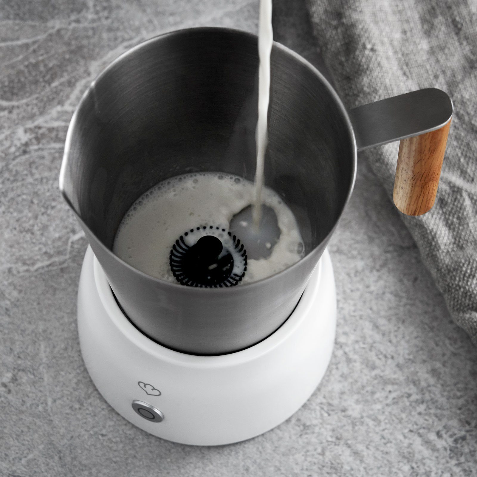 Milch, die in eine Aufschäummaschine mit Holzgriff gegossen wird. Die Maschine ist weiß.