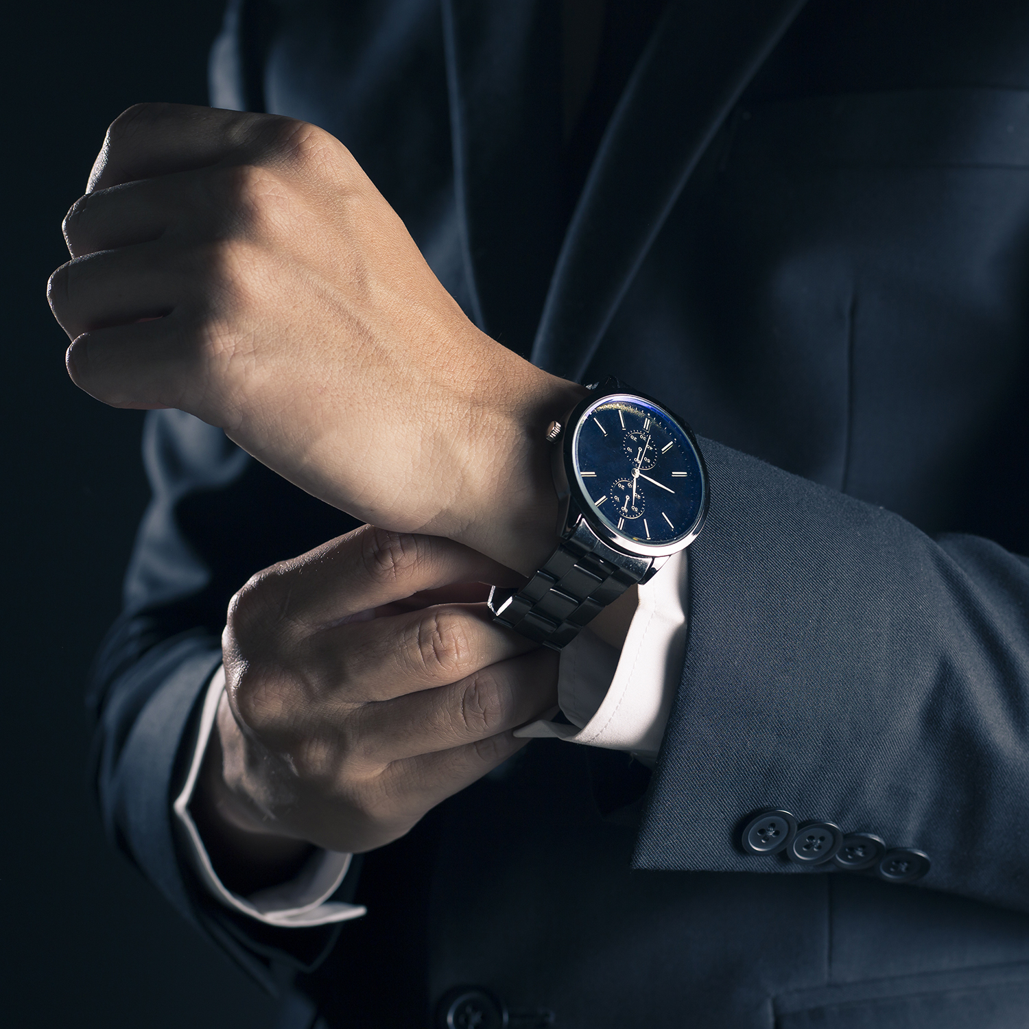 Ein Mann im Anzug justiert seine Armbanduhr. Die Uhr hat ein schwarzes Armband und ein dunkelblaues Zifferblatt.
