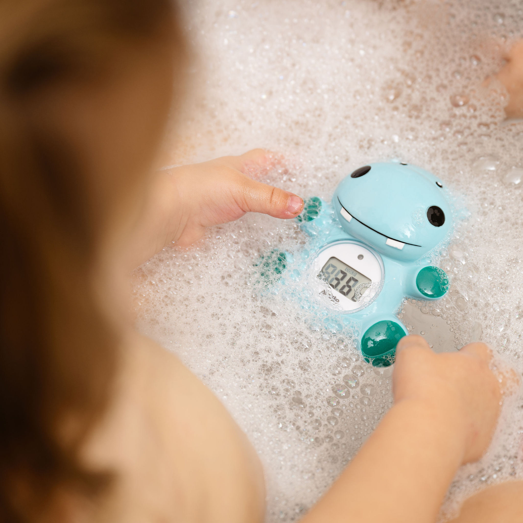 Dziecięce ręce trzymają niebieski termometr do kąpieli w wannie z bąbelkami. Termometr pokazuje temp.