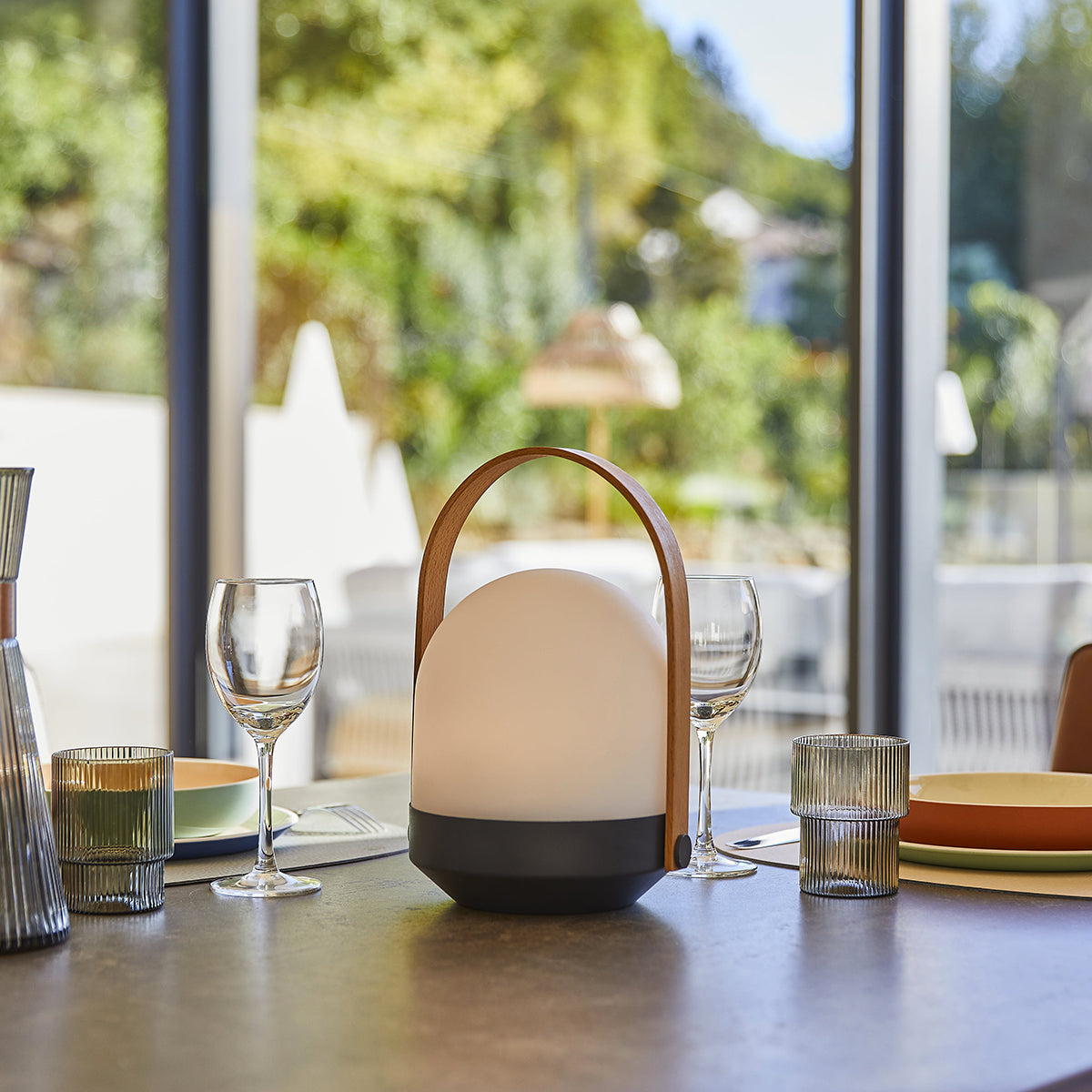 Eine Tischdekoration mit einer weißen Lampe, Weingläsern, Tellern und einem grünen Ausblick.