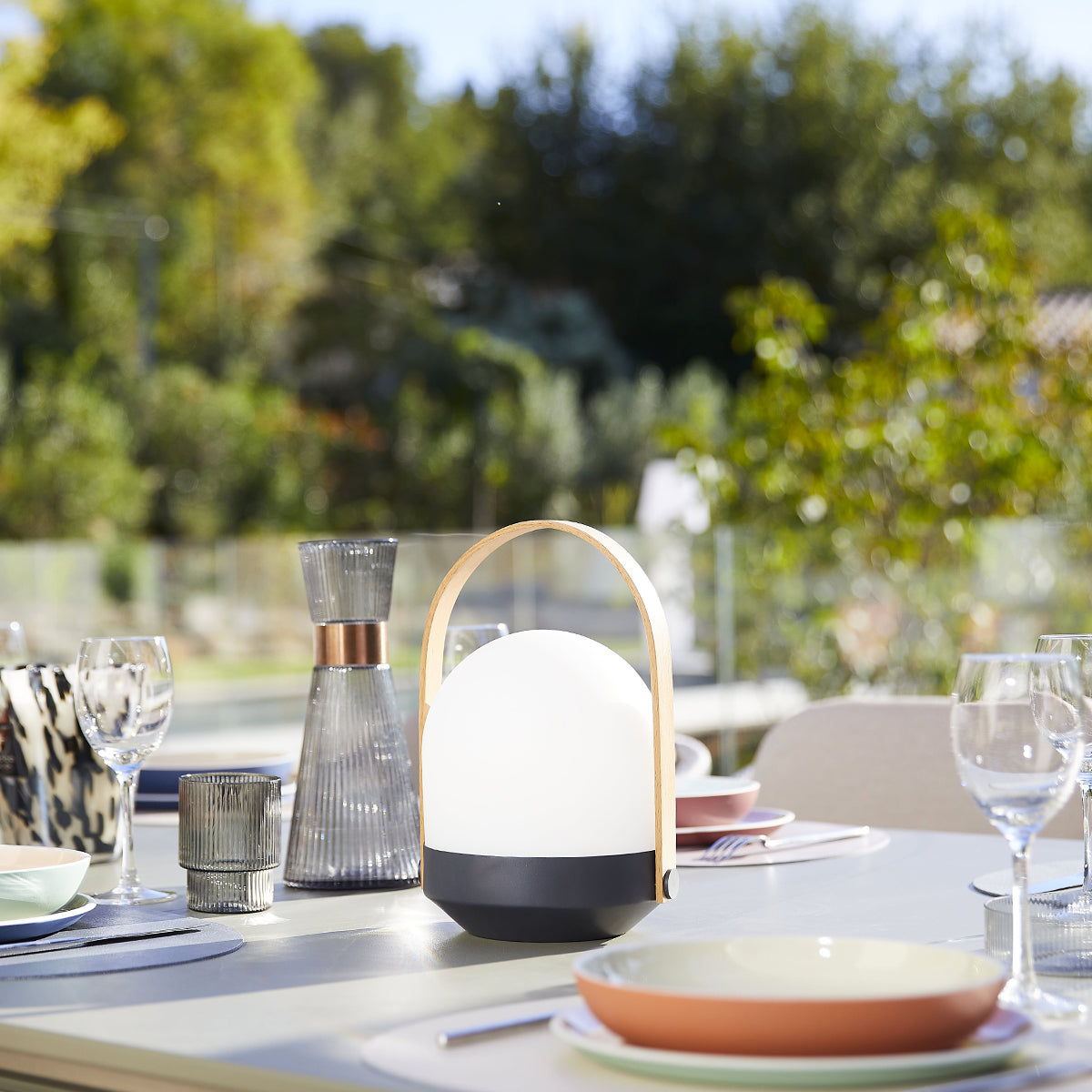 Szene im Freien mit einer Lampe und Gläsern auf einem Tisch. Der Hintergrund ist mit Bäumen verschwommen. Sonniger Tag.