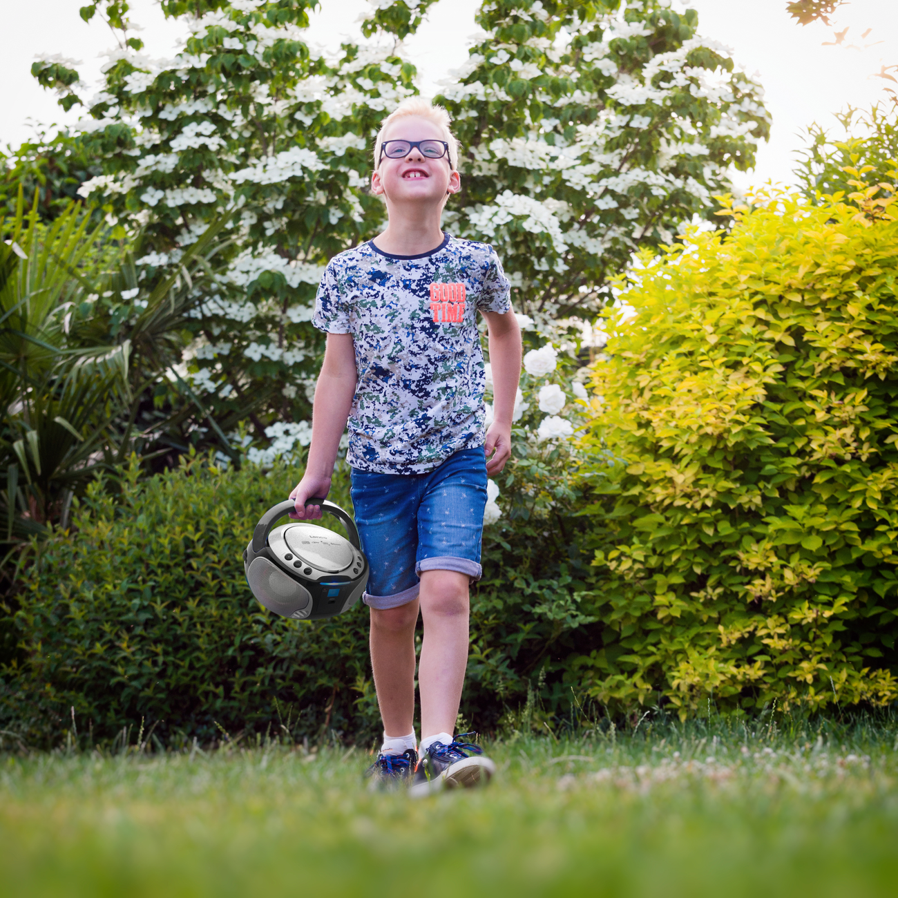 Boy walking on grass carrying a portable speaker, smiling. Background with flowers.