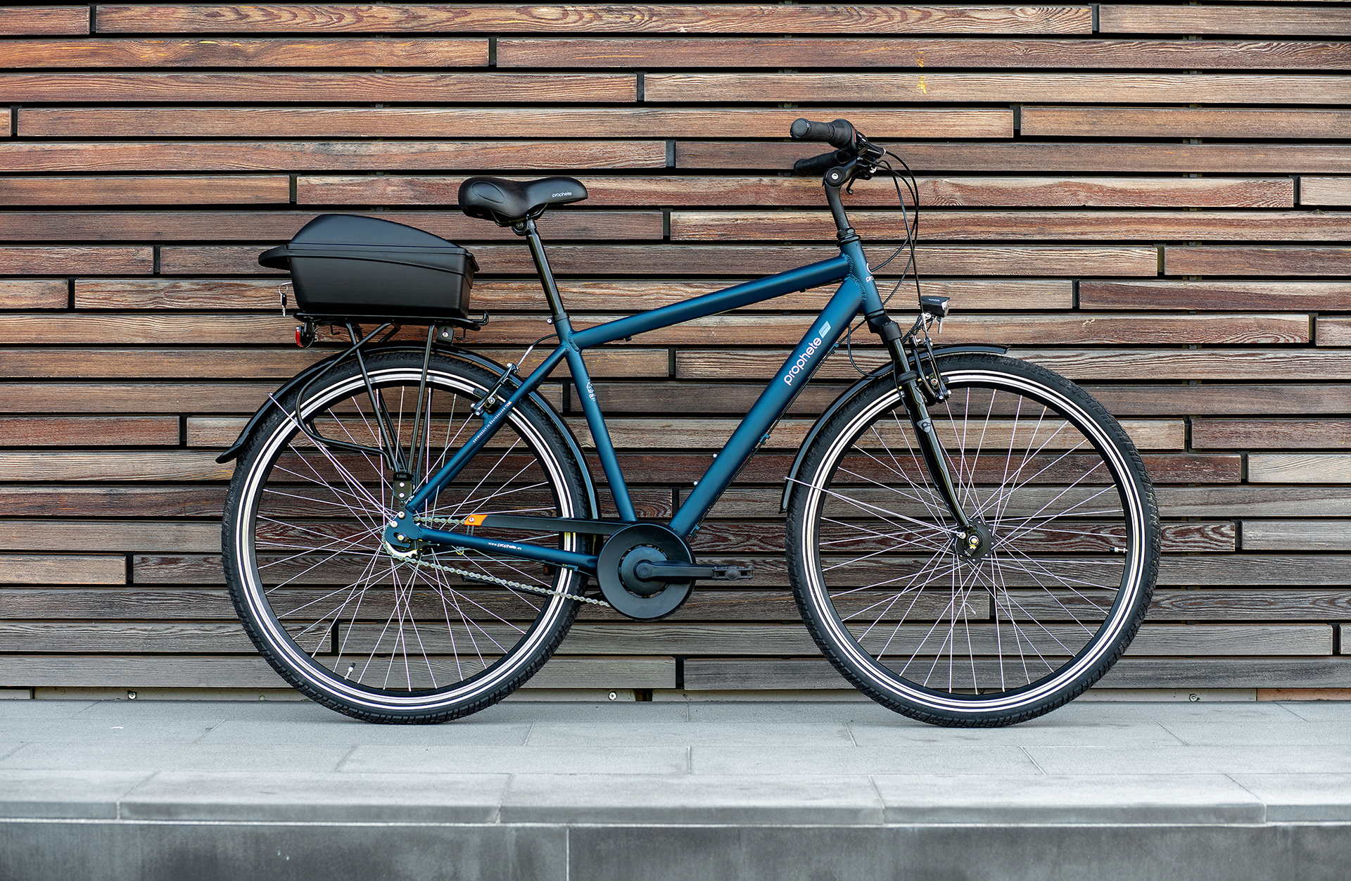 Ein blaues Fahrrad mit einer schwarzen Gepäckbox, geparkt vor einer holzgetäfelten Wand.