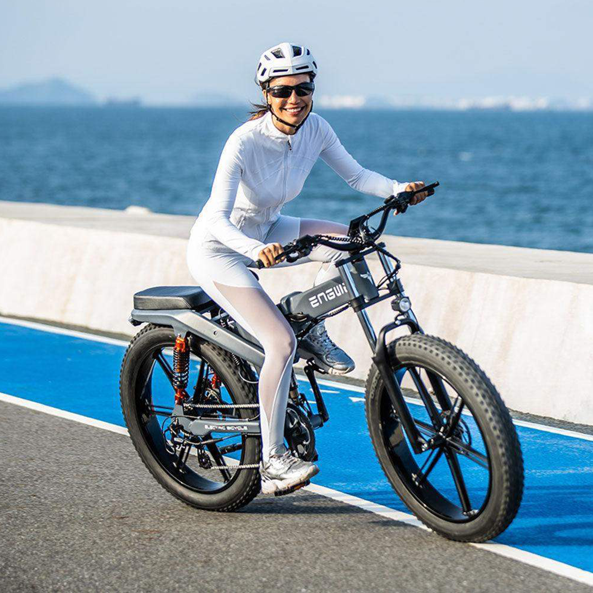 Kobieta jadąca szarym rowerem elektrycznym po drodze obok morza.