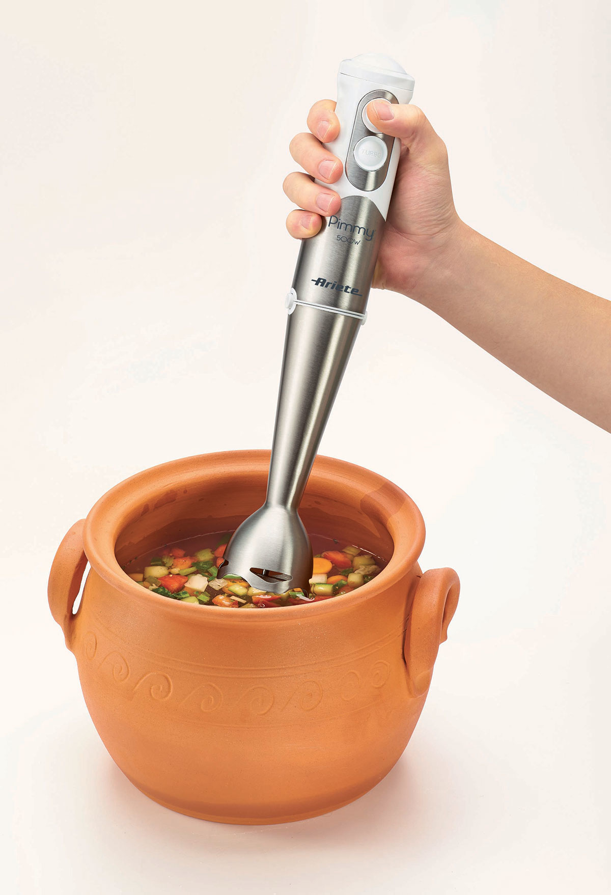 Una mano che tiene un frullatore a immersione argento e bianco in una pentola di terracotta con zuppa.