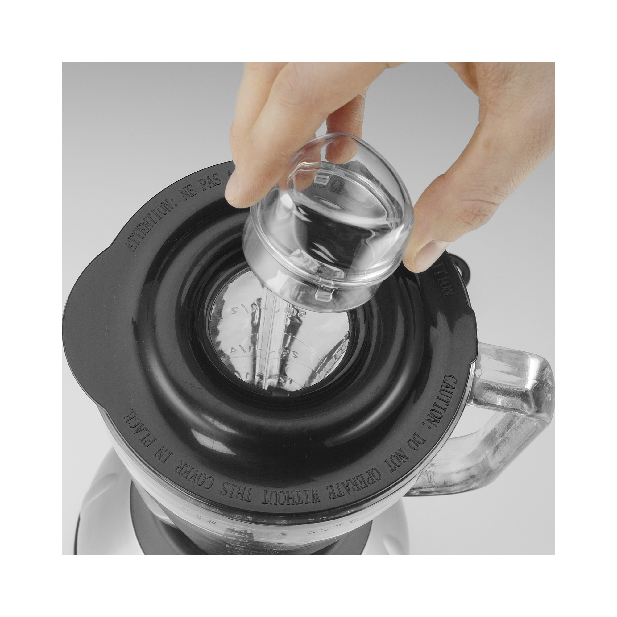 Hand gießt Wasser aus einem kleinen, durchsichtigen Behälter in einen Mixer.