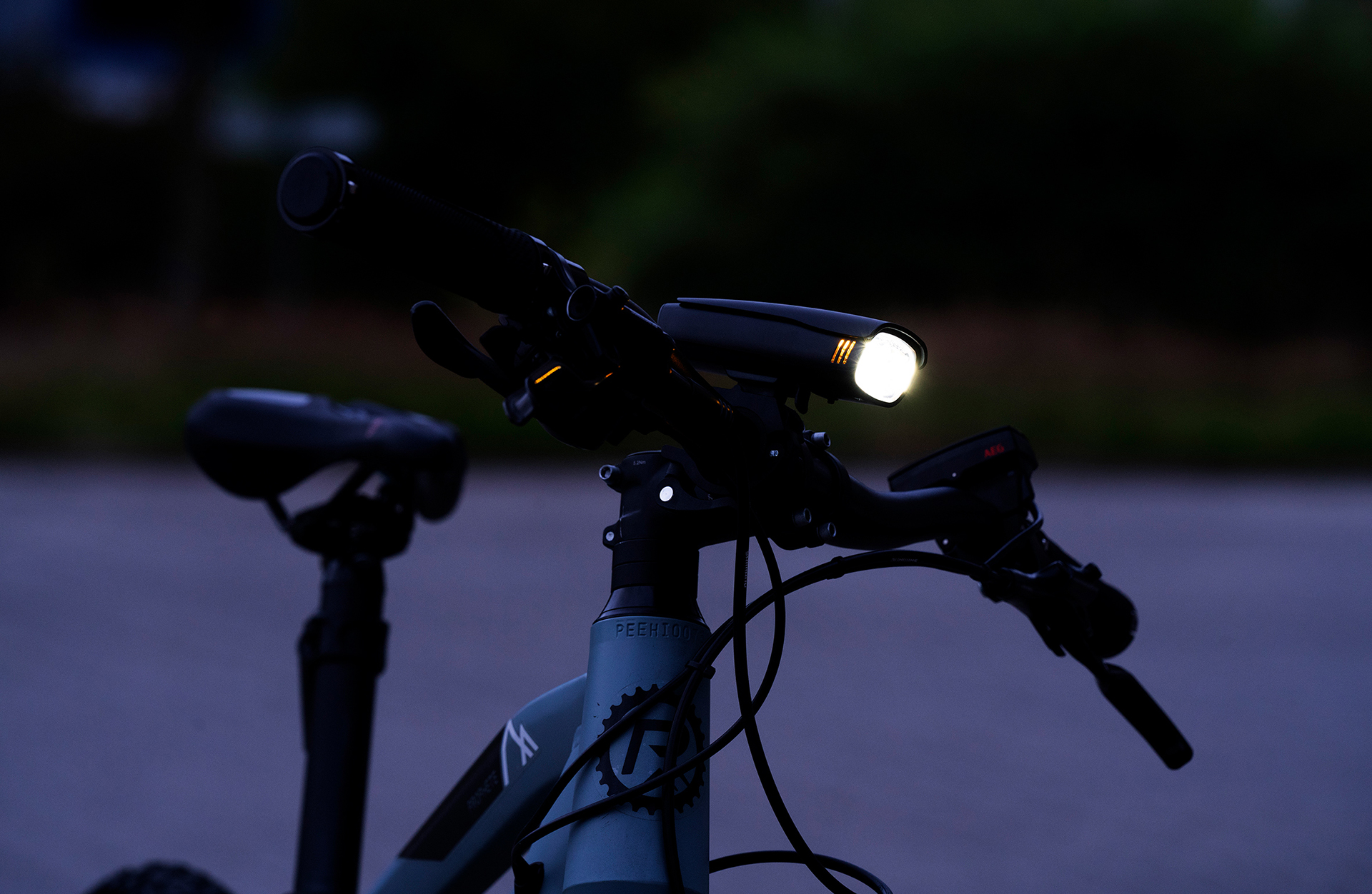 Fahrradlenker mit eingeschaltetem Scheinwerfer, in dunkler Umgebung. Fahrradsattel im Hintergrund sichtbar.