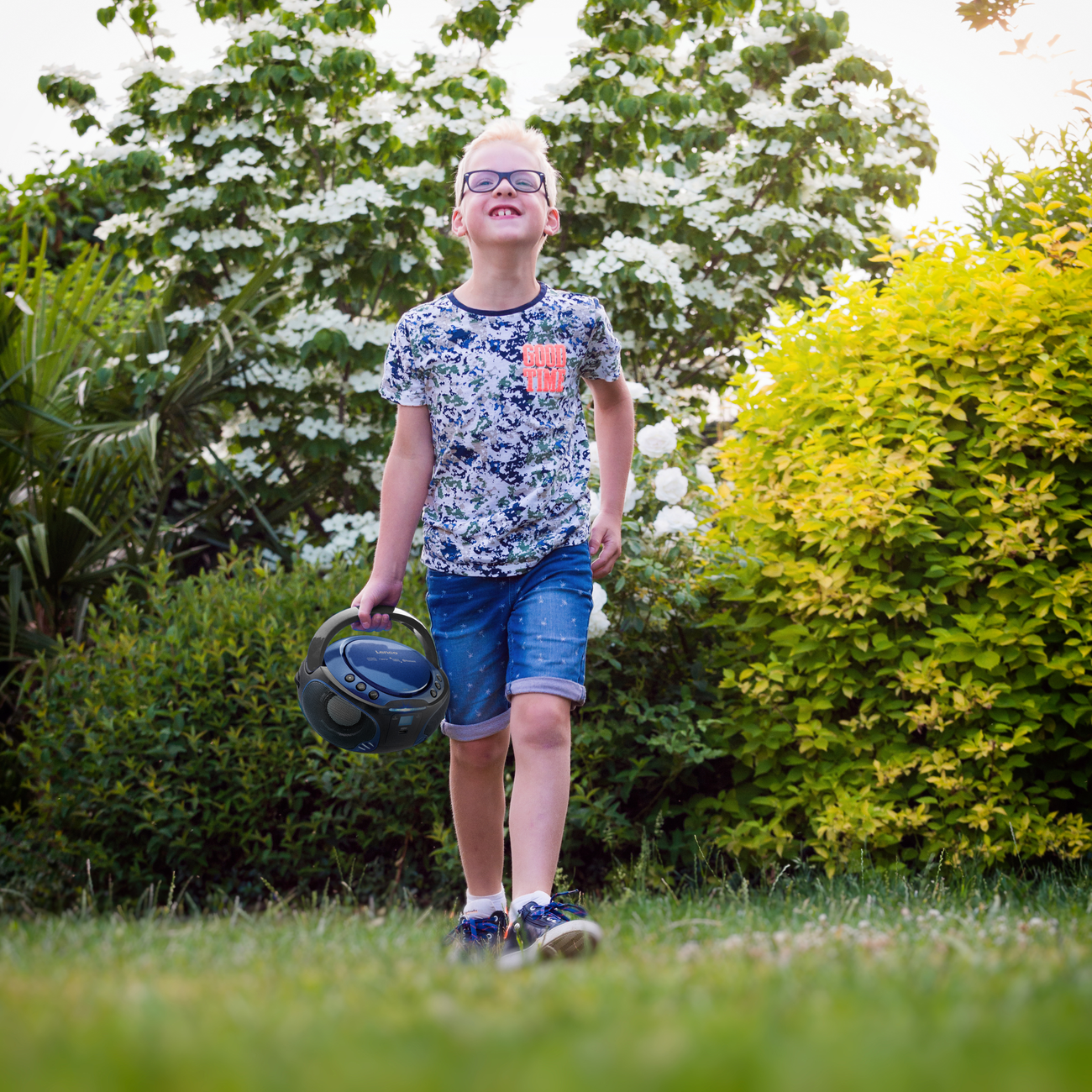 Boy with glasses walks holding a boombox in a garden. Green grass, white flowers, and green bushes.