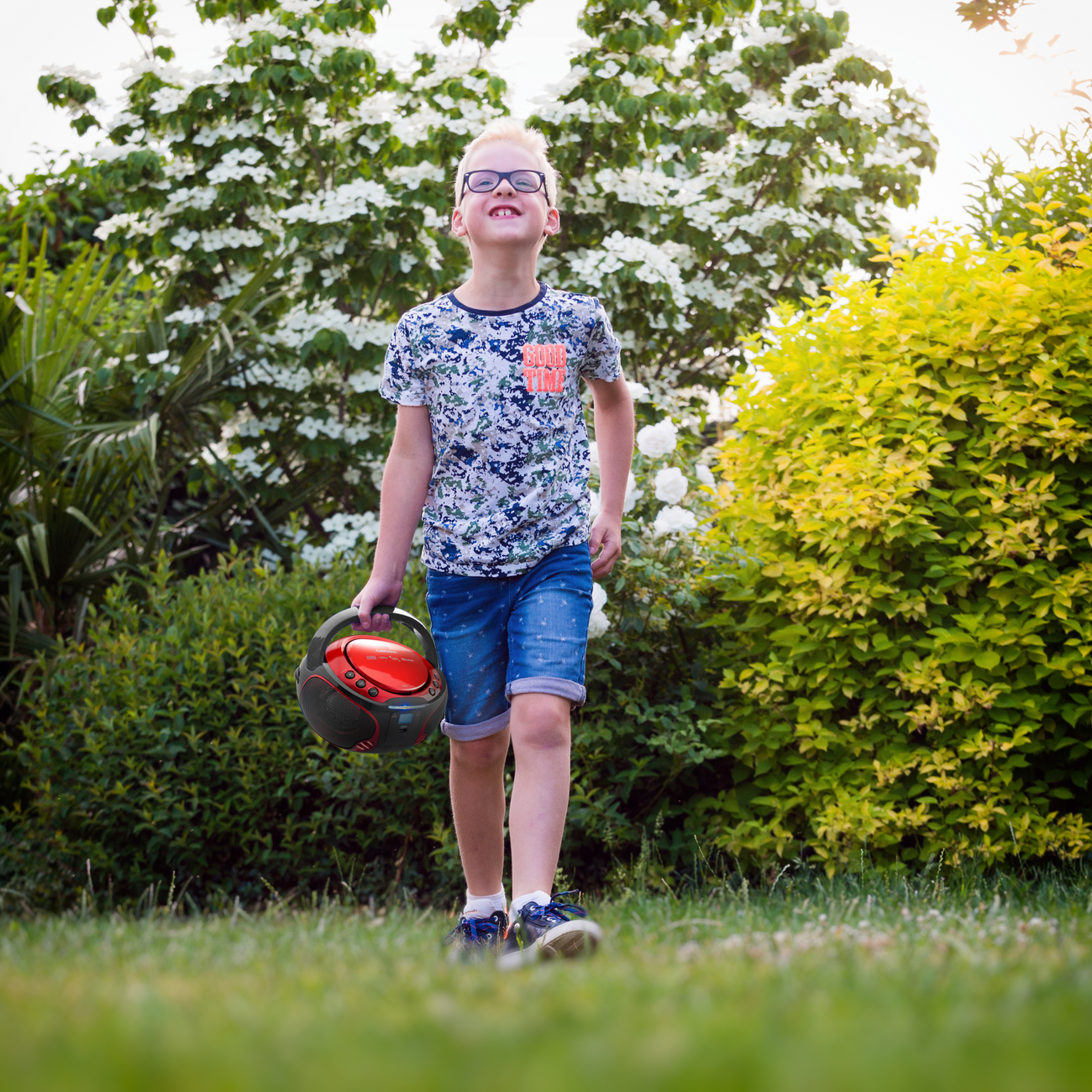 Boy walking on grass, holding a red and black boombox. Green trees and white flowers in background.
