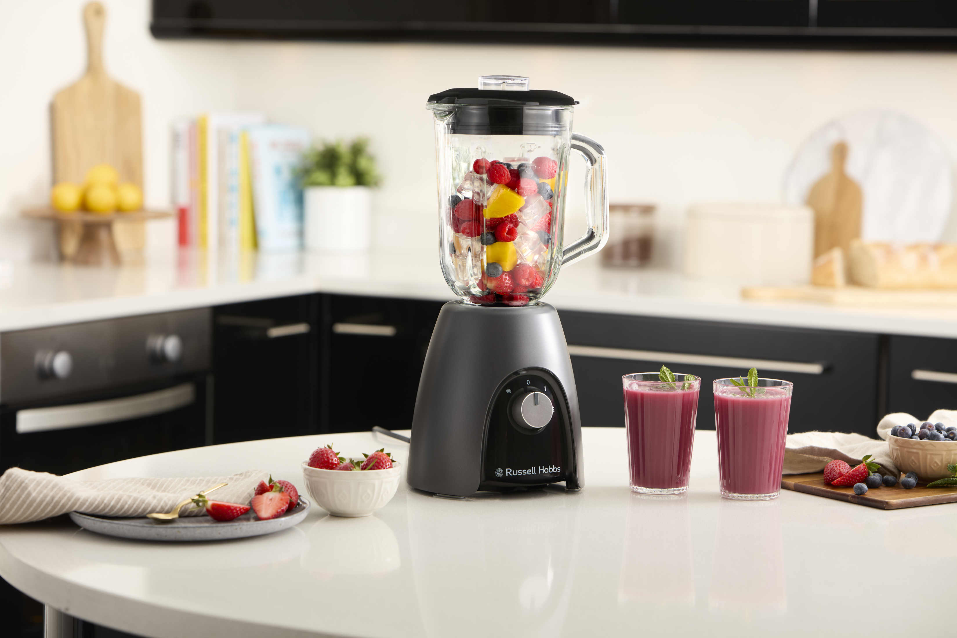 Mixer mit Früchten und zwei Gläsern Smoothie auf einem weißen Tisch in der Küche.