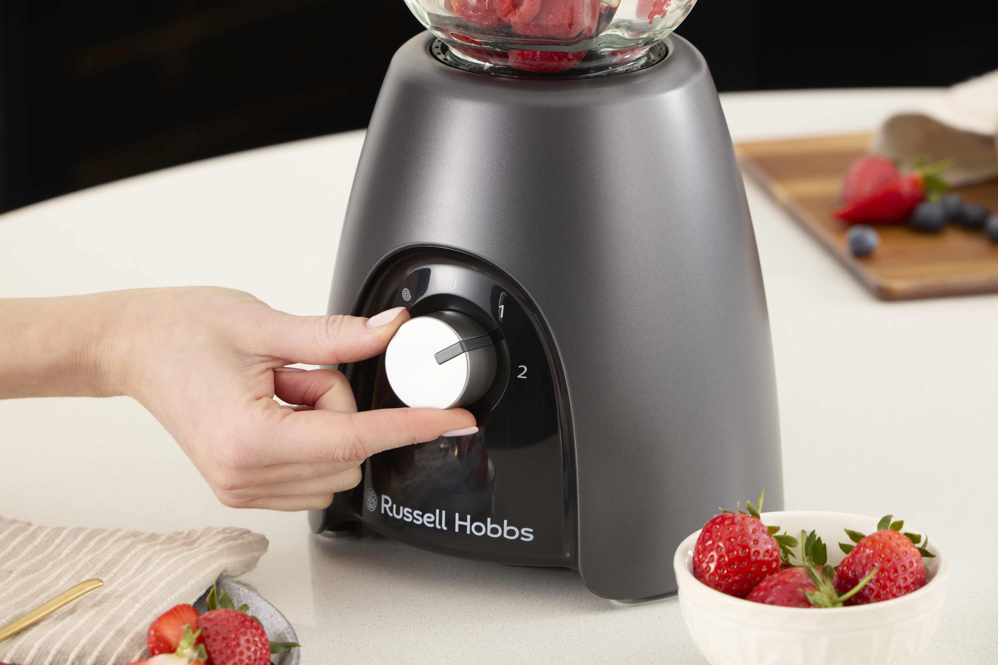 Eine Hand dreht einen Mixer-Regler mit Erdbeeren in einer Schüssel und auf einem Tisch.
