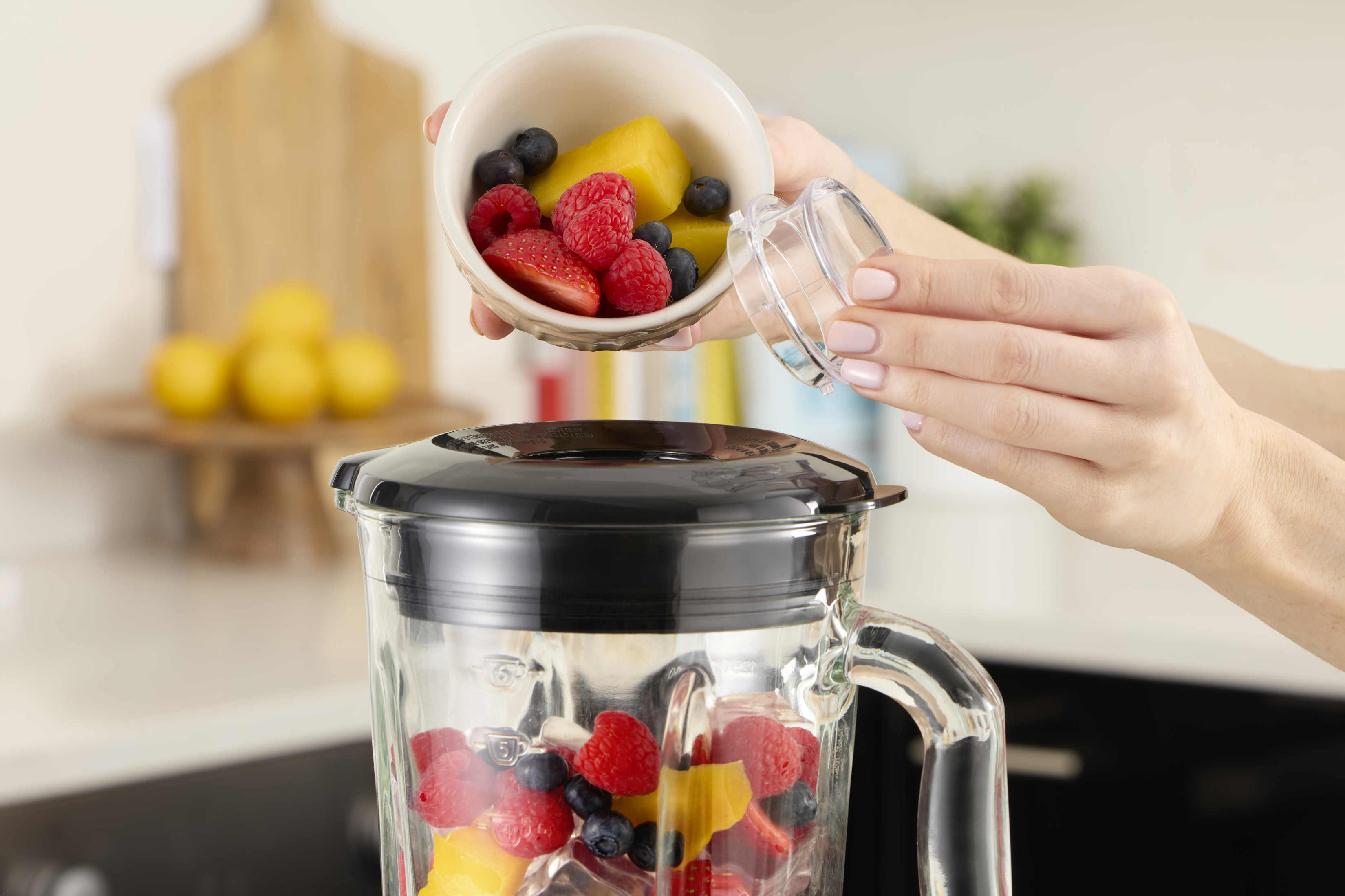 Eine Frau gießt Früchte in einen Mixer. Der Mixer hat einen Glasbehälter mit schwarzem Deckel und enthält Früchte.
