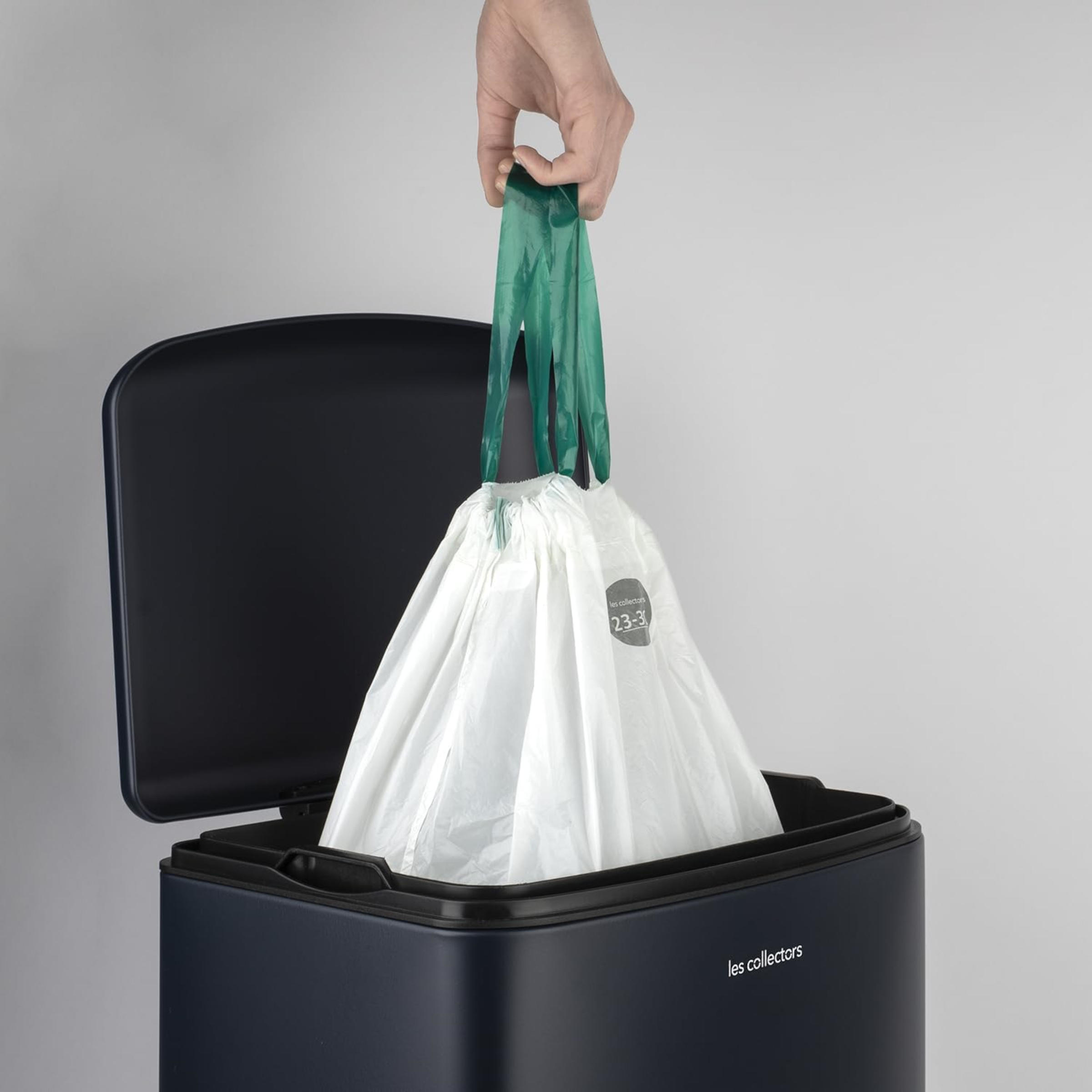 Una mano sostiene una bolsa de basura verde con cordón sobre un cubo de basura azul oscuro.
