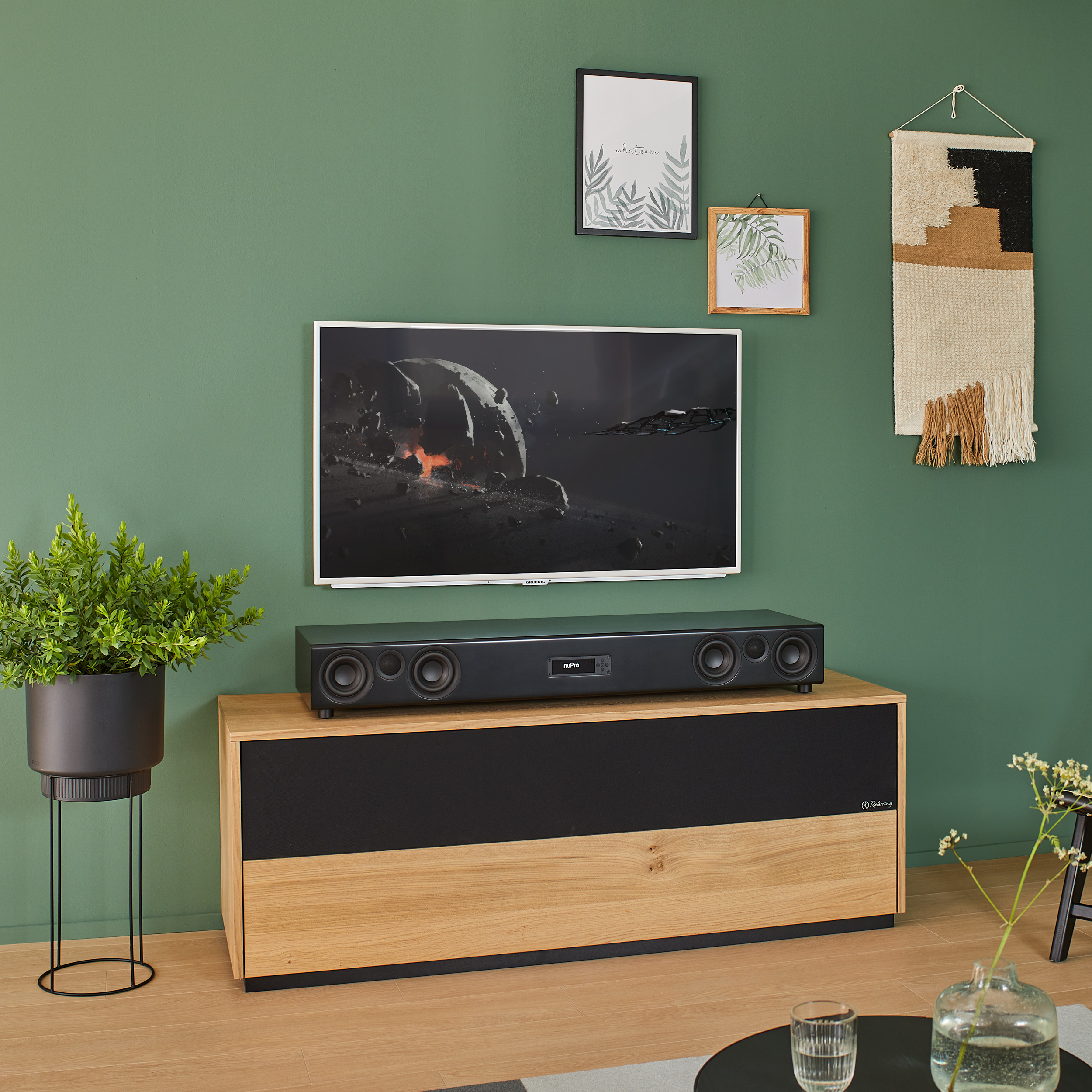 Ein Fernseher auf einem Holzschrank, mit einer Soundbar, Pflanzen und gerahmten Bildern an einer grünen Wand.