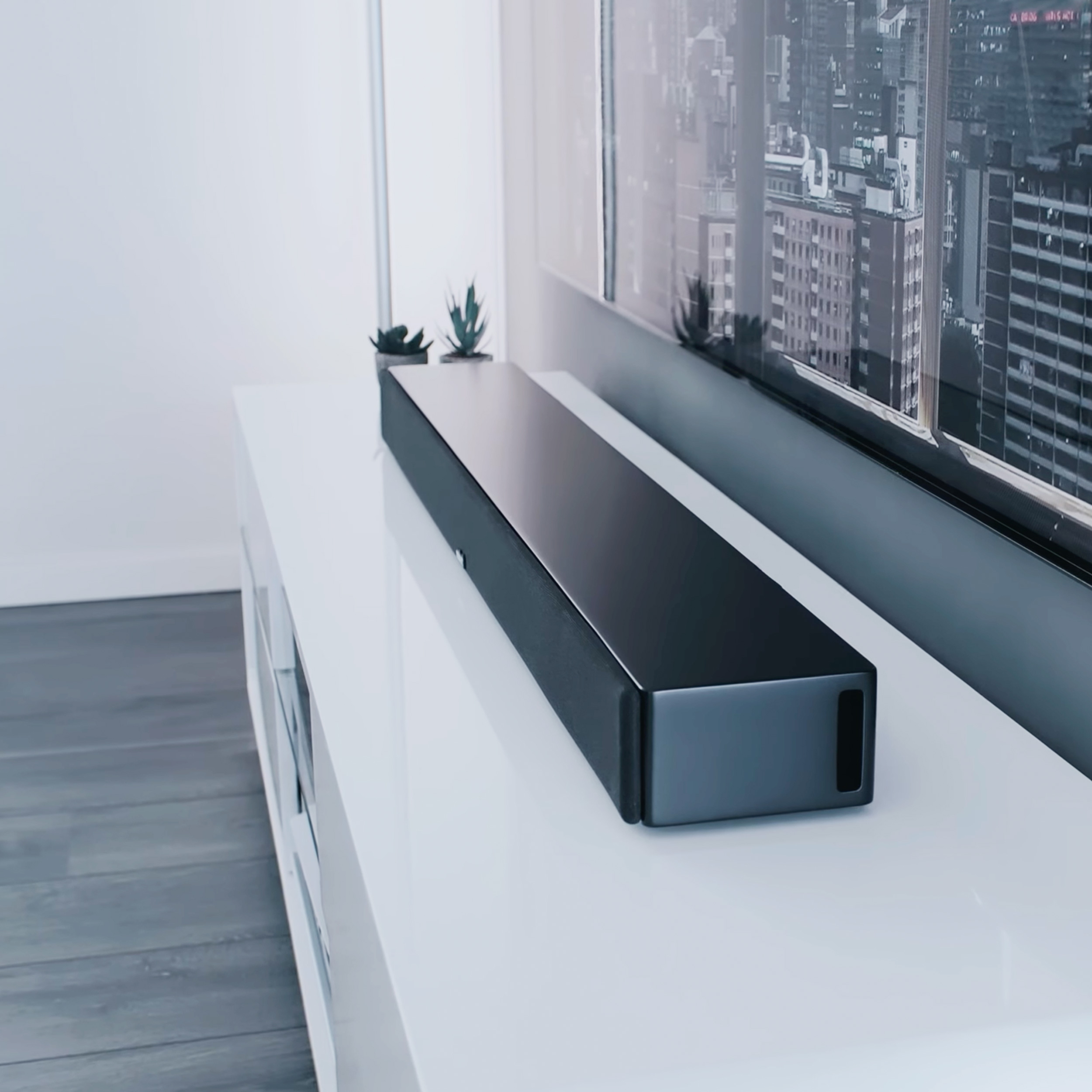 Schwarze Soundbar auf weißem Schrank. Ein Fernseher und Stadtbild im Hintergrund.