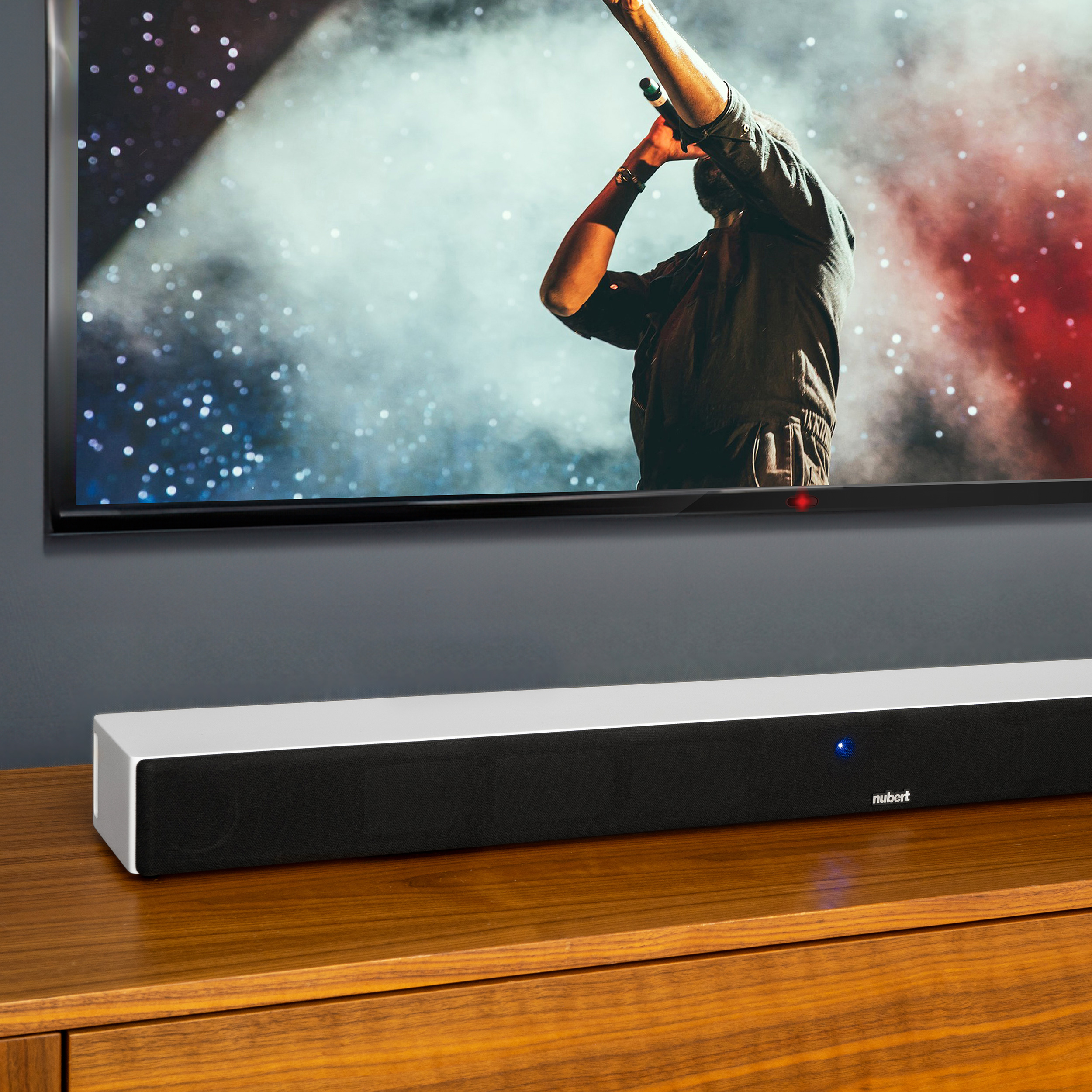 Eine Soundbar steht auf einer Holzoberfläche unter einem Fernseher, der ein Konzert zeigt.