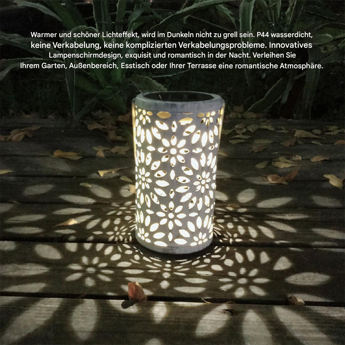 Eine zylindrische Gartenlaterne aus Metall mit floralen Aussparungen, die Lichtmuster auf eine Holzoberfläche wirft.