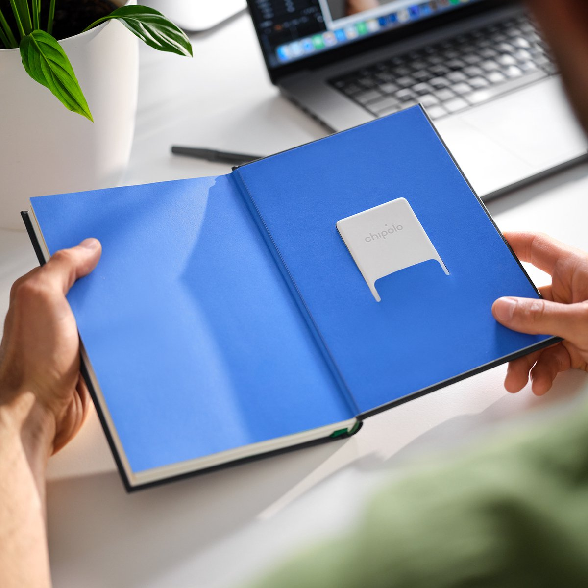 Mani aprono un libro blu con un dispositivo Chipo bianco. Un laptop sullo sfondo.