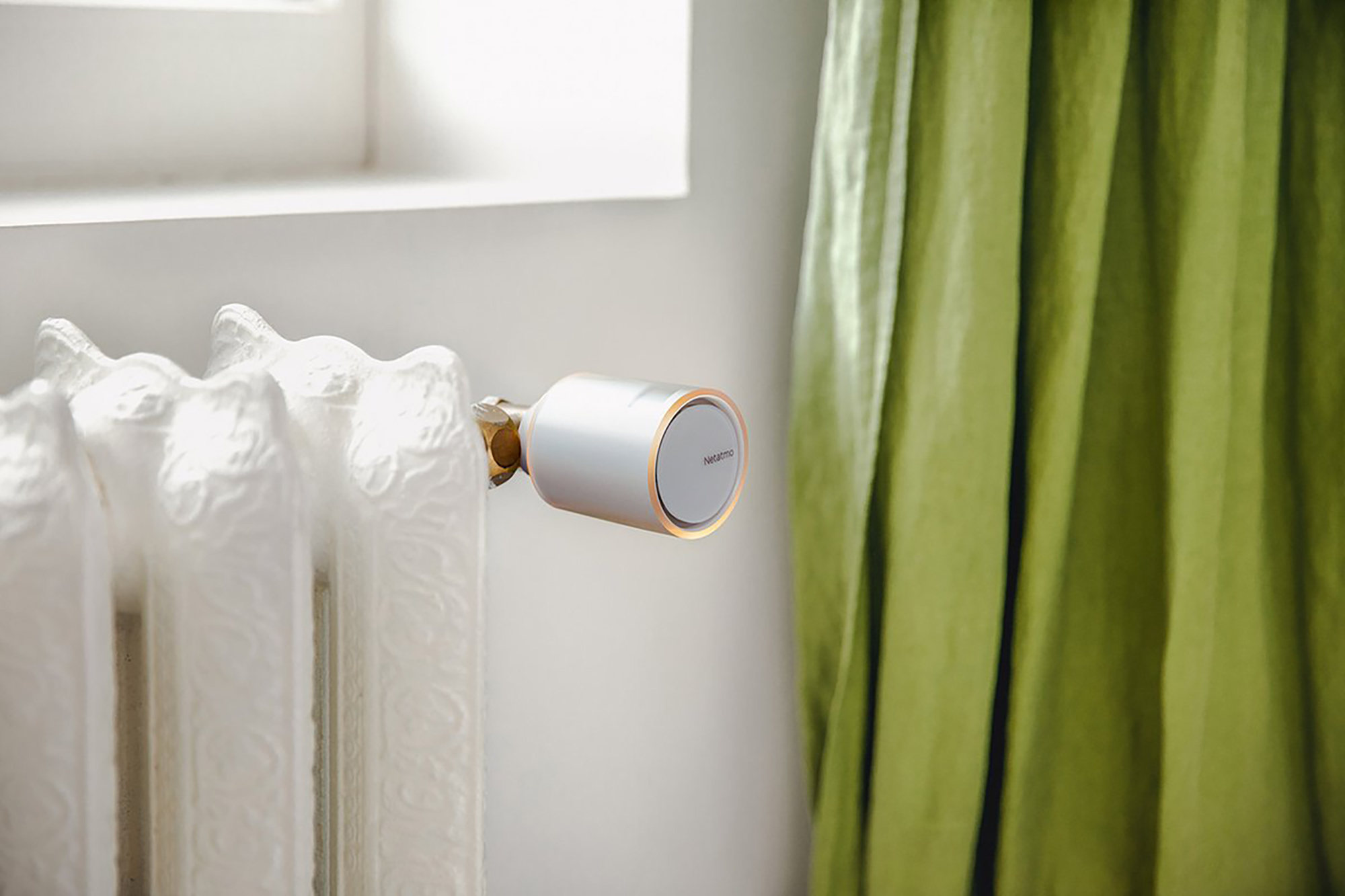 A smart thermostat installed on a white radiator next to a green curtain.