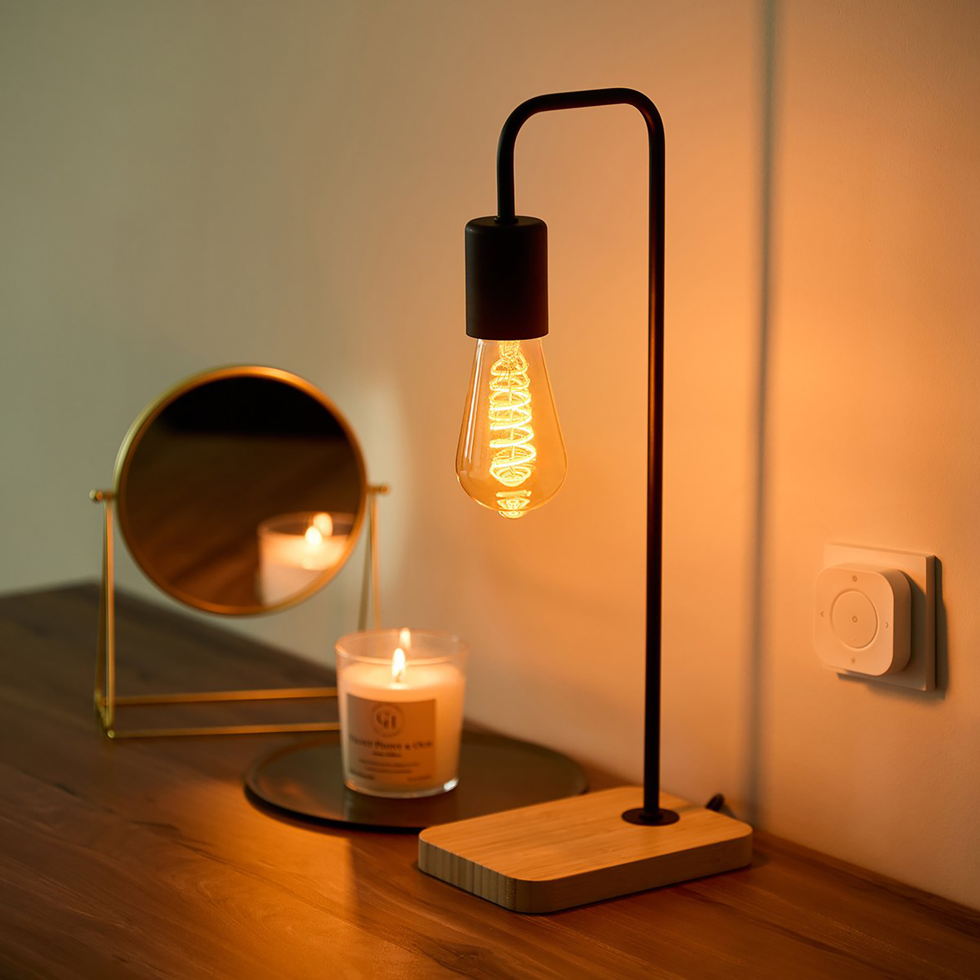 A lamp with a vintage bulb on a wooden desk. A mirror and candle are also present.