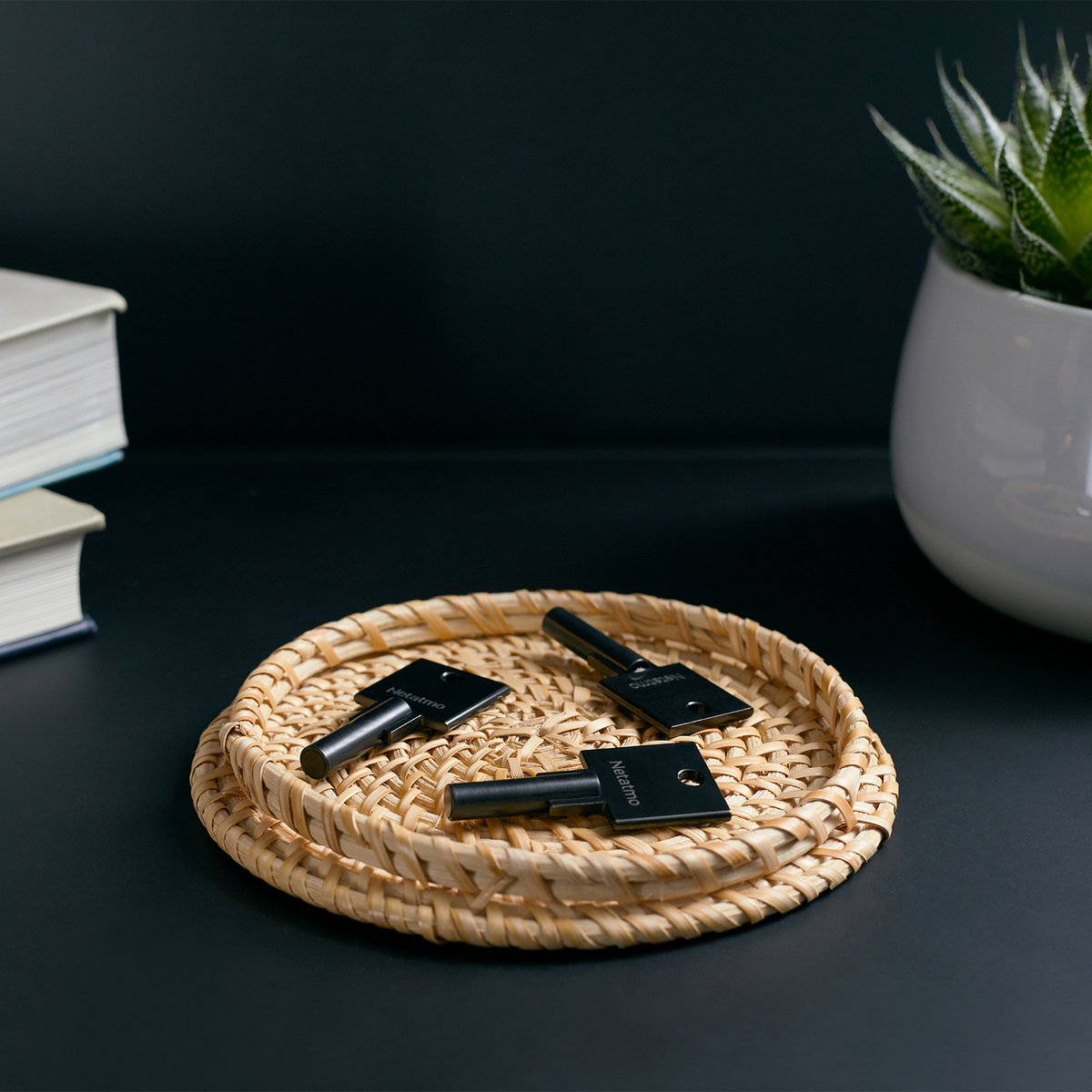 Three black Netatmo keys are in a woven basket on a dark surface, with a plant and books nearby.