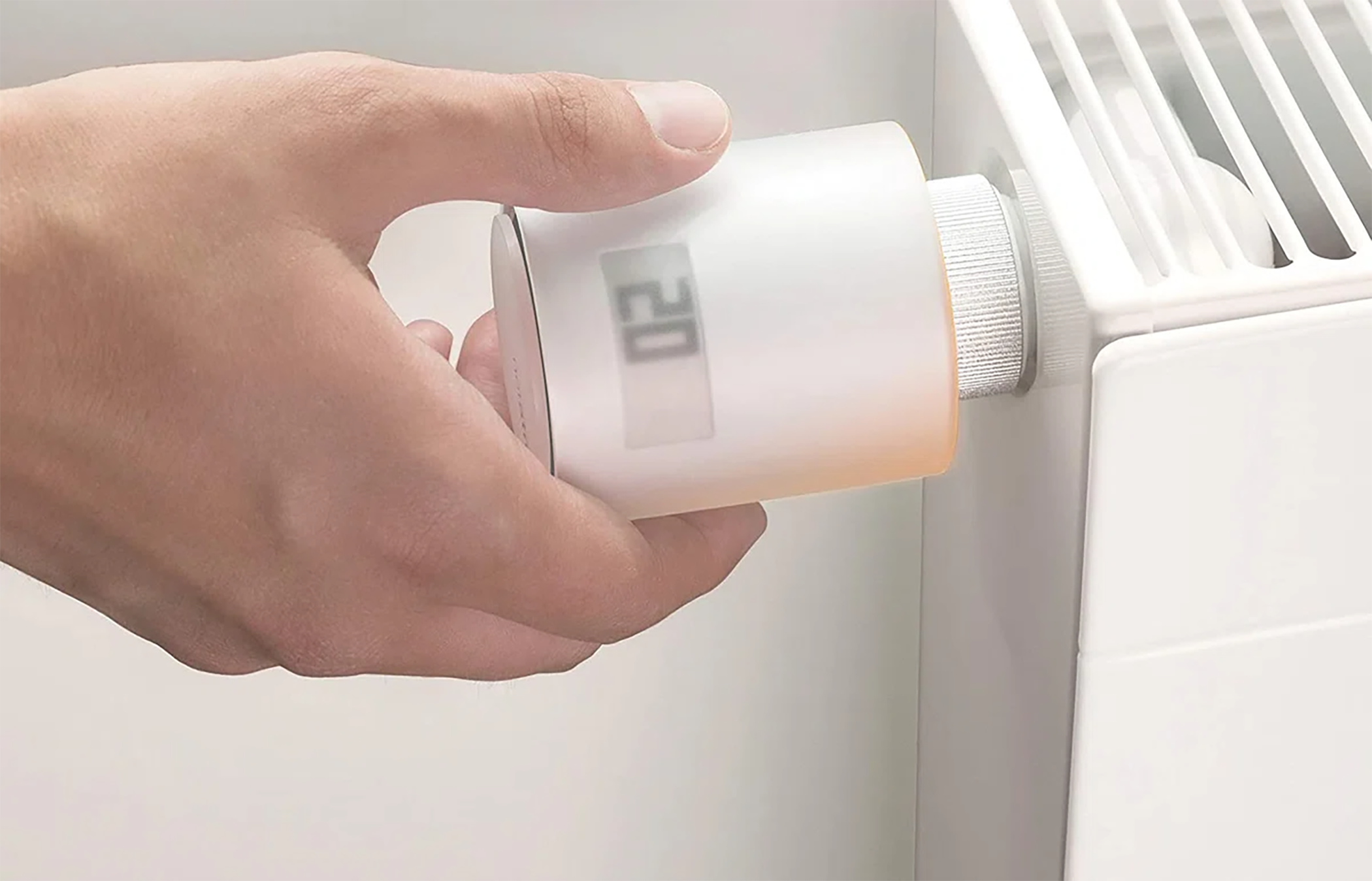 Hand adjusting a white smart thermostat on a radiator, showing the temperature.