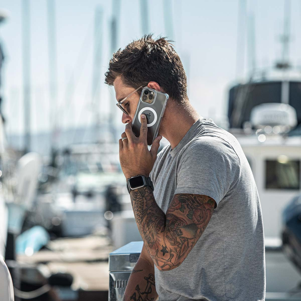 Een man met tatoeages belt met een telefoon in een doorzichtige hoes. De achtergrond toont boten en de zee.