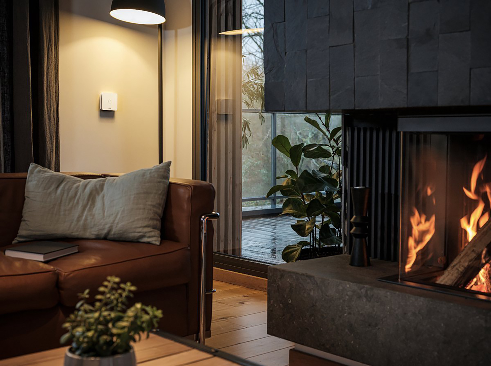 Gezellige woonkamer met open haard, bruine leren bank en lamp. Een lichtschakelaar aan de muur.