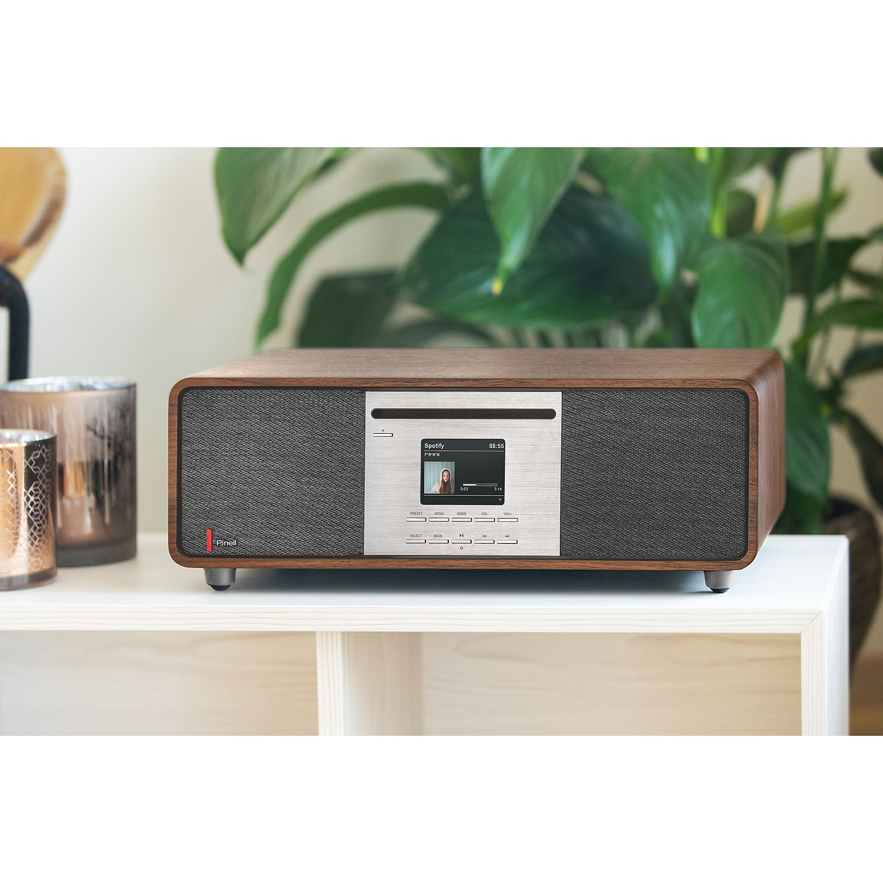 Ein Holzradio mit einem Display. Das Radio steht auf einer weißen Oberfläche. Im Hintergrund befinden sich dekorative Gegenstände.