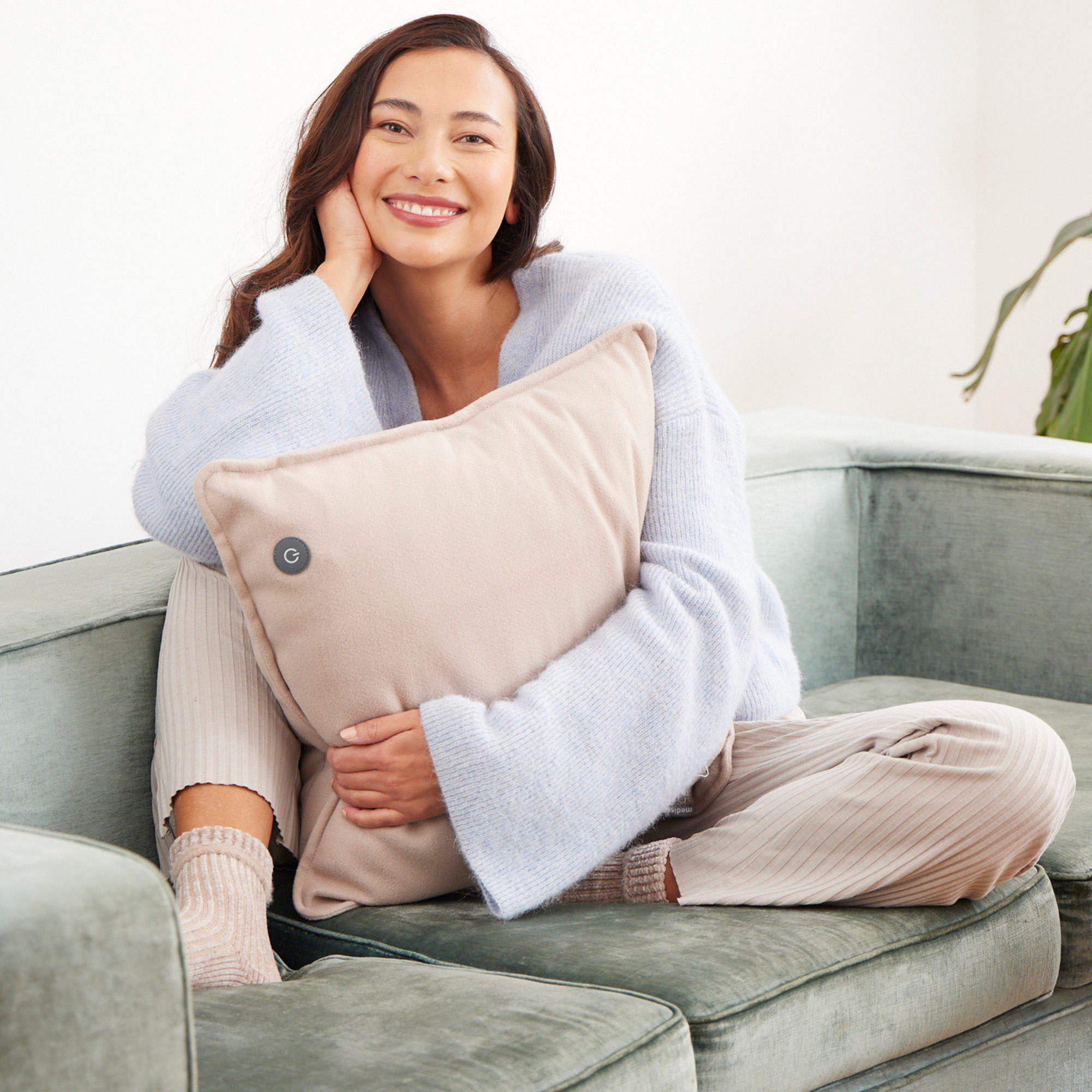Eine Frau lächelt und hält ein beiges Kissen, sitzt auf einem grünen Sofa.