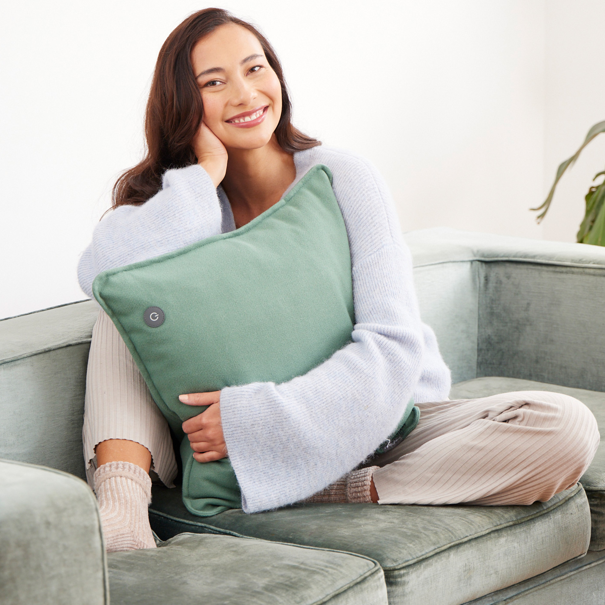 Una mujer sonriendo, sosteniendo una almohada verde en un sofá verde, con un suéter azul claro.