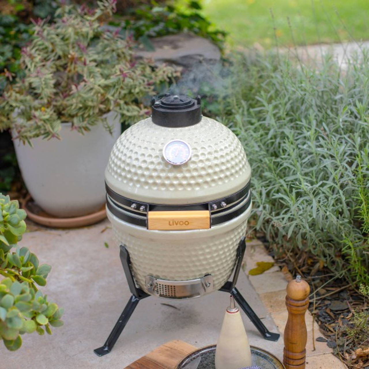 Ein beiger Keramikgrill mit Holzgriff steht auf einer Terrasse, Rauch steigt auf. Pflanzen und eine Flasche im Hintergrund.