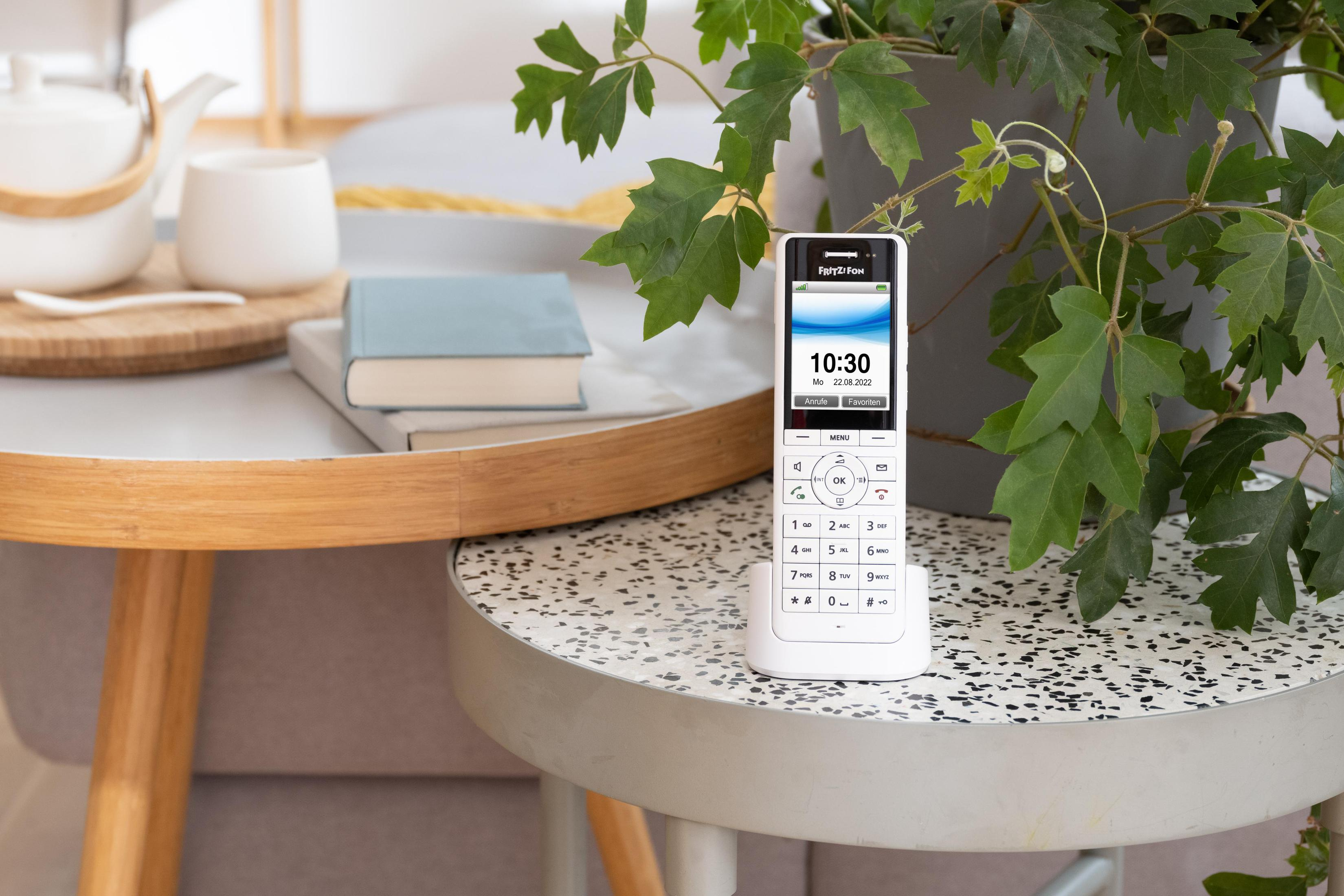 Un téléphone sans fil blanc sur une table avec un livre, une plante et une télécommande, avec une horloge affichée.