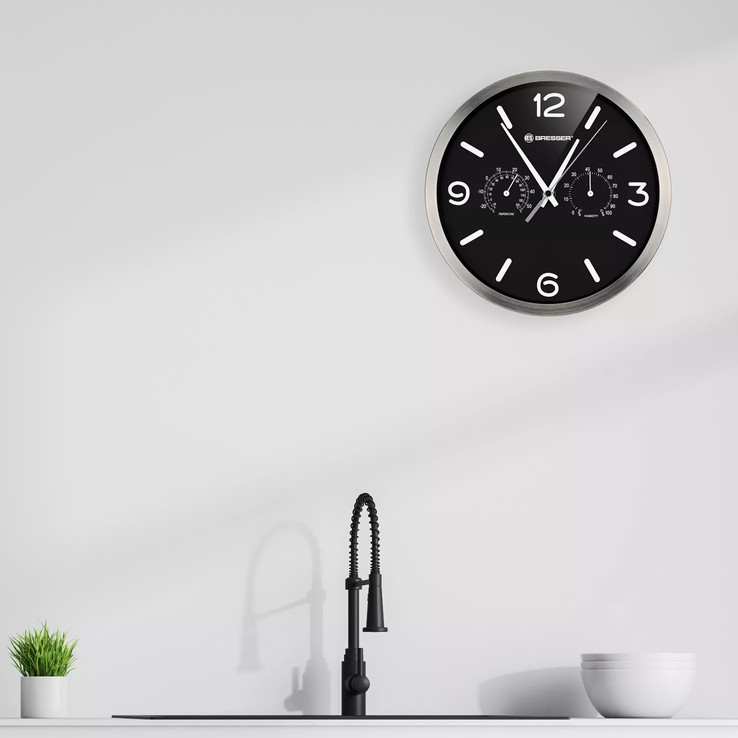 Schwarze und silberne Uhr mit Ziffern und Zifferblättern. Schwarzer Wasserhahn und Pflanze davor.