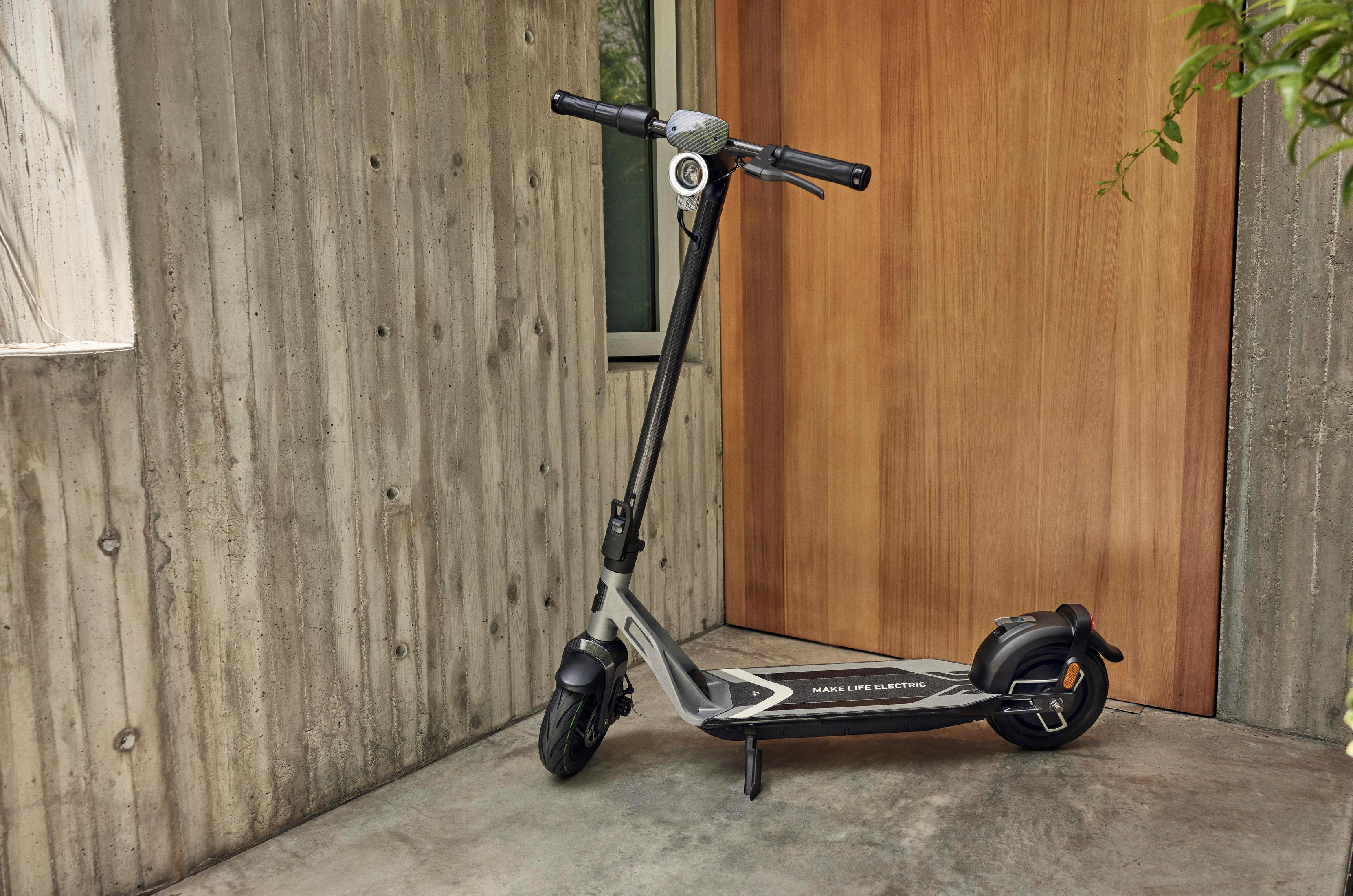 Elektroroller geparkt an einer Betonmauer und Holztür. Roller ist schwarz und grau.