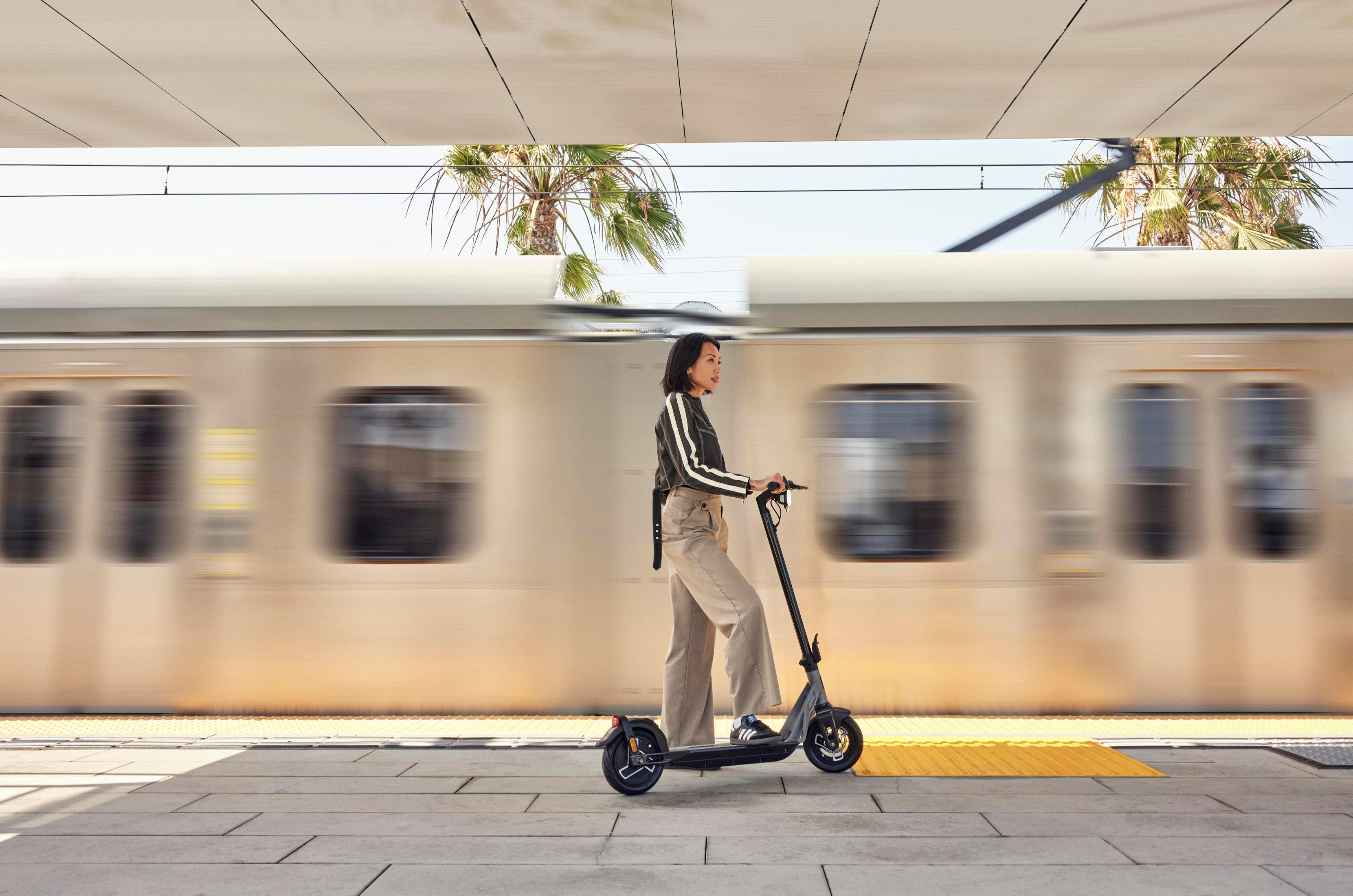 Frau fährt Roller am Bahnhof, Zug im Hintergrund, Palmen sind sichtbar.