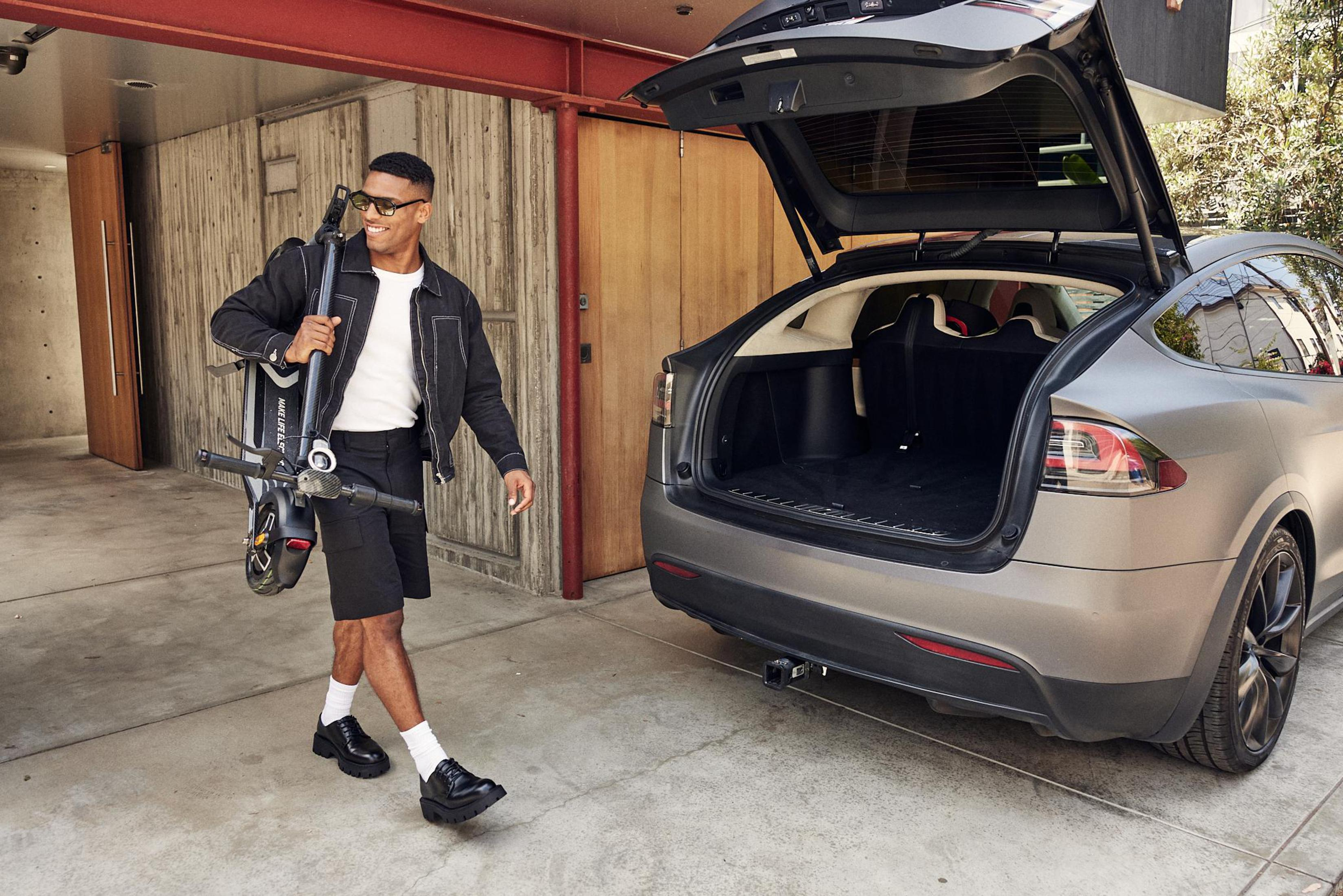 Ein Mann in schwarzen Shorts, Jacke und Sonnenbrille geht auf einen grauen SUV zu, mit einem Roller.