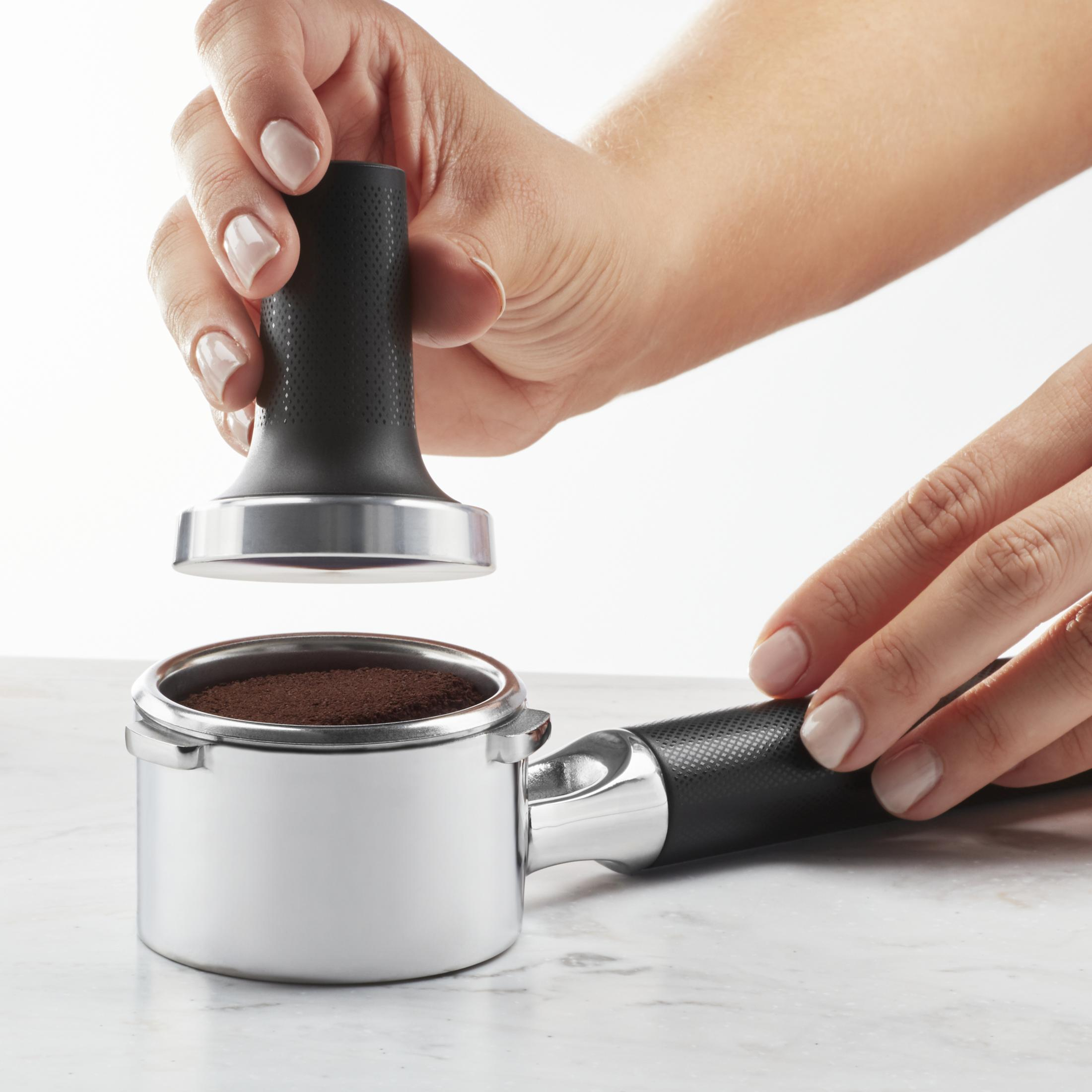 Hände einer Person, die einen Tamper verwenden, um Kaffeepulver in einen Metall-Siebträger zu pressen.