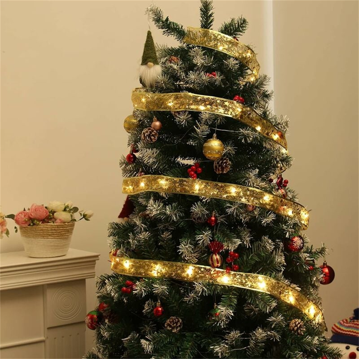 Weihnachtsbaum, geschmückt mit goldenem Band, Lichtern und Ornamenten. Ein Wichtel thront auf der Spitze. Blumen im Hintergrund.