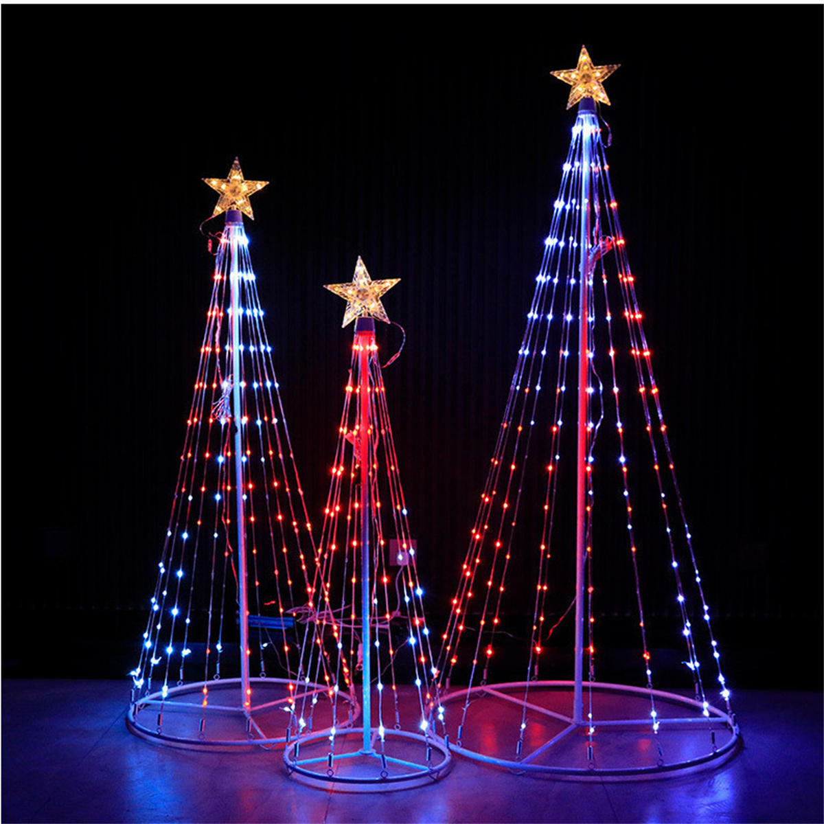 Drei beleuchtete Weihnachtsbäume mit Sternaufsätzen. Bäume sind weiß mit roten und blauen Lichtern, auf dunklem Grund.