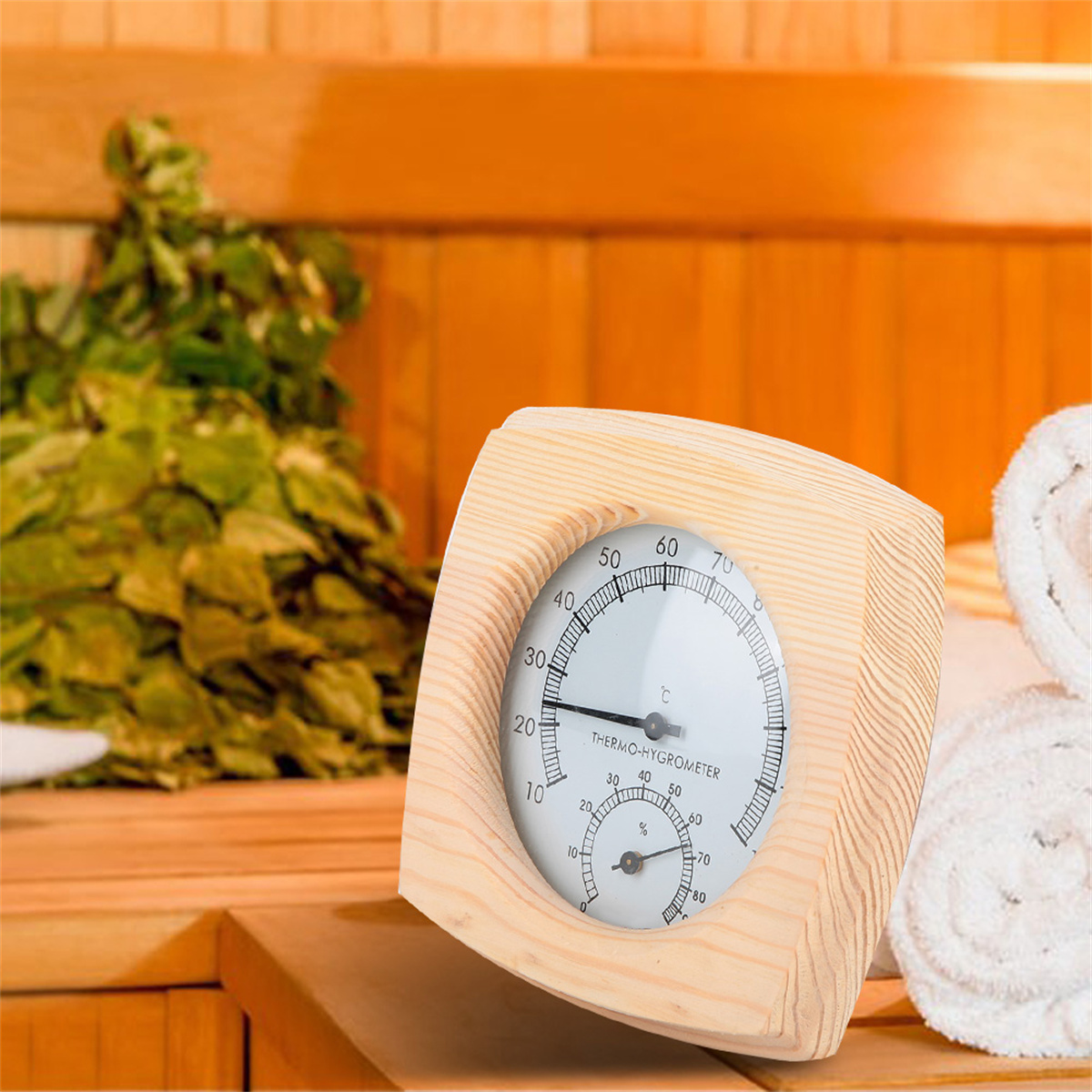 Ein hölzernes Sauna-Thermometer mit rundem Zifferblatt, auf einer Holzbank. Grüne Blätter und weiße Handtücher im Hintergrund.