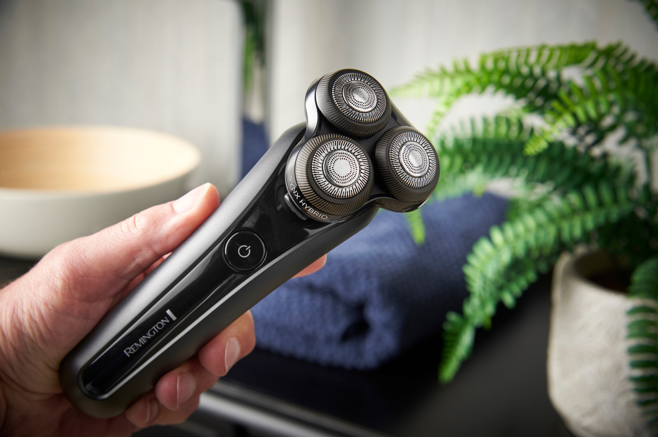 Eine Hand hält einen schwarzen Remington-Rasierer. Grüne Pflanze, blaues Handtuch und eine weiße Schüssel im Hintergrund.